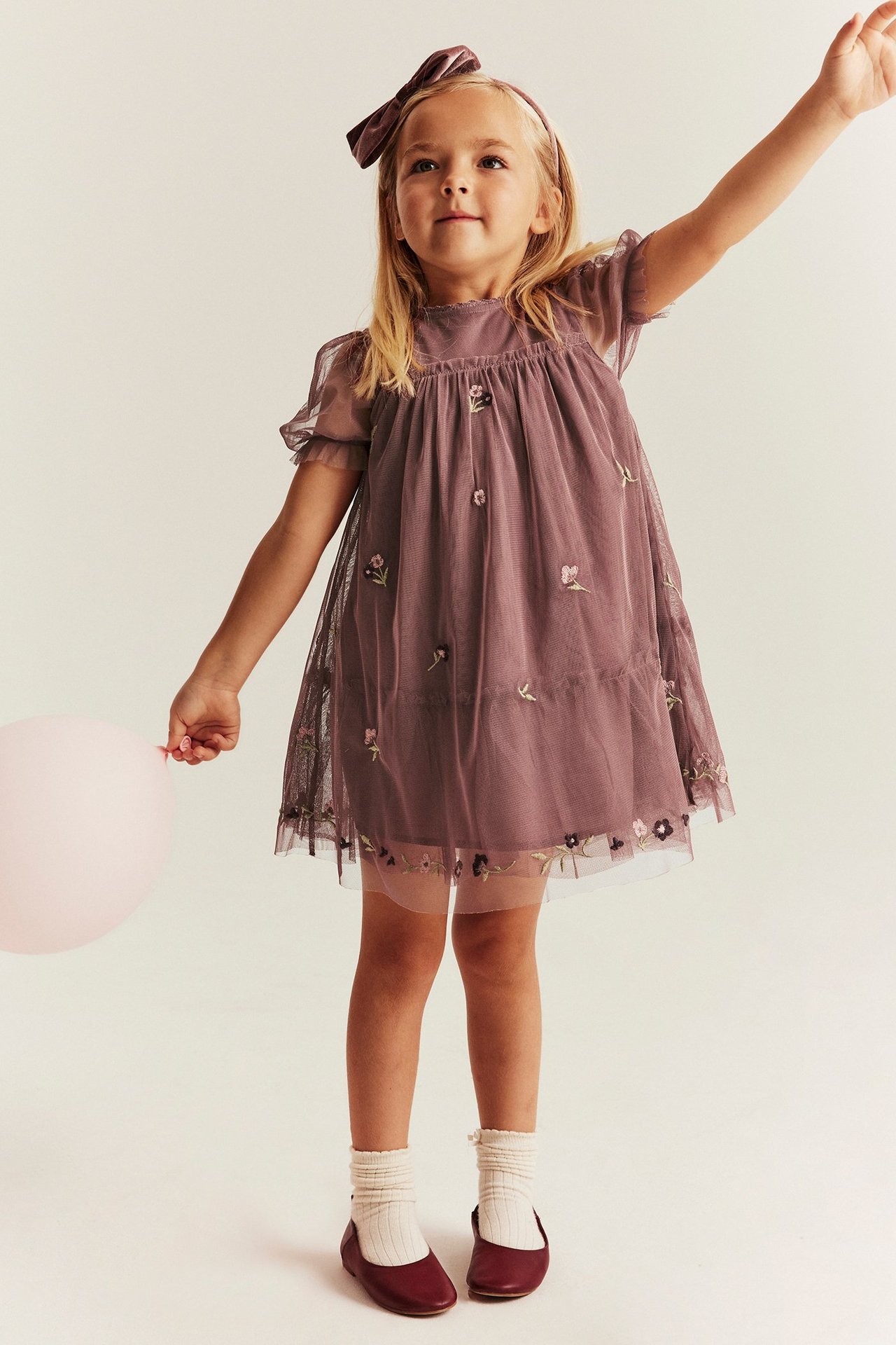Front view: Kid in a dark pink embroidered mesh dress with puff sleeves, velvet headband, white socks, holding a balloon.