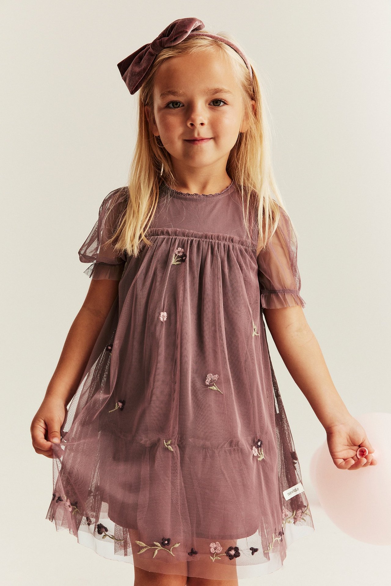 Kid in dark pink embroidered mesh dress, puff sleeves, velvet bow headband, holding a pink balloon.