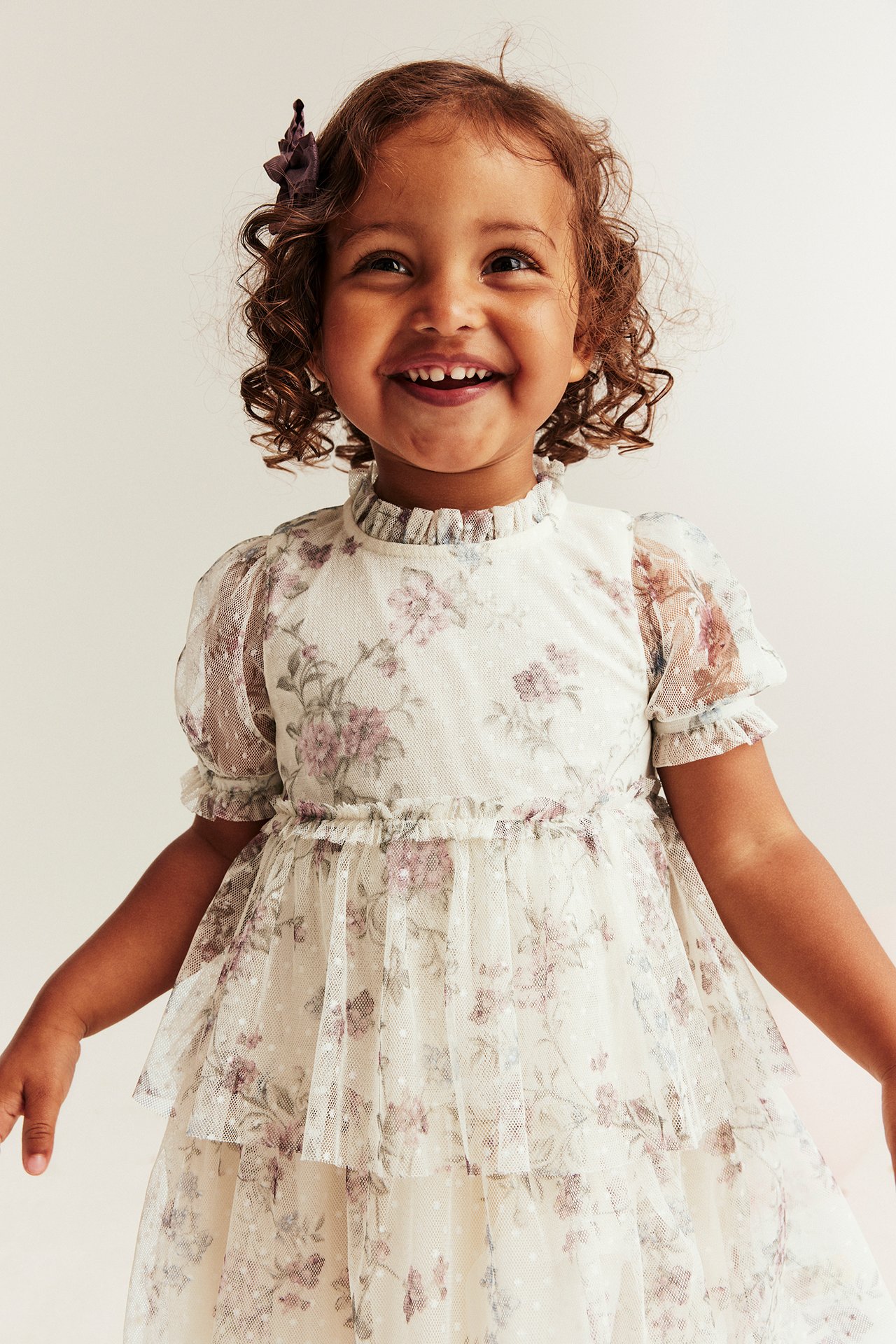 Smiling kid wearing a light beige floral mesh dress with ruffled collar, puff sleeves, and tiered skirt.