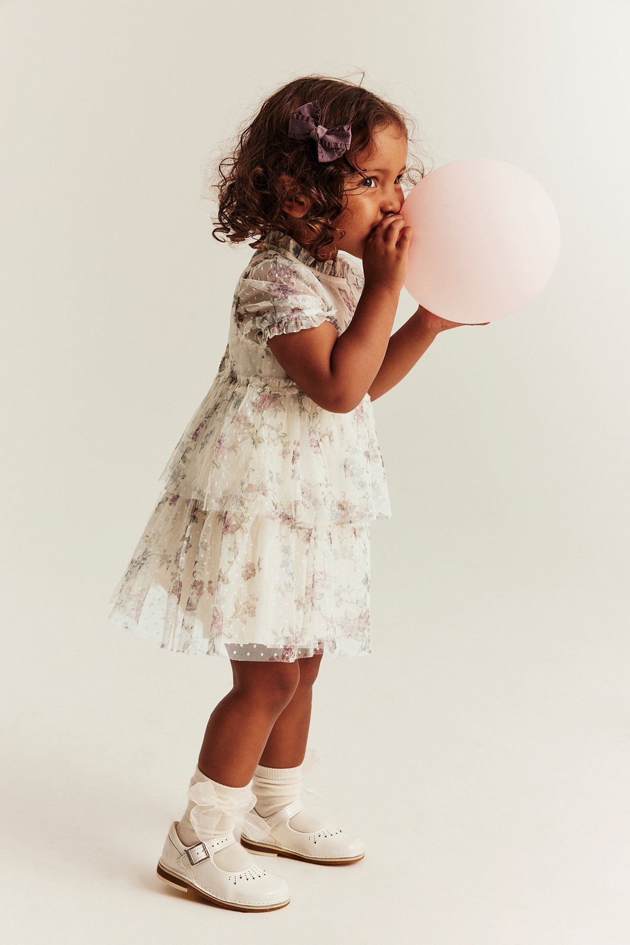 Side view of a kid in a light beige floral mesh tiered dress with puff sleeves, blowing a pink balloon.