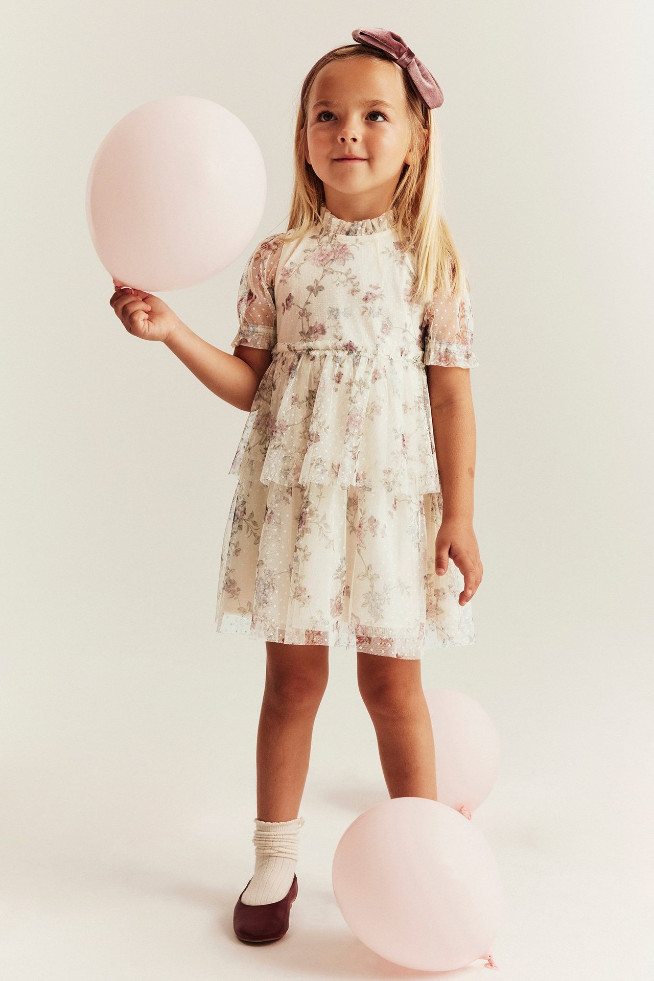 Girl in light beige floral mesh dress with ruffle skirt and puff sleeves, holding a pink balloon.