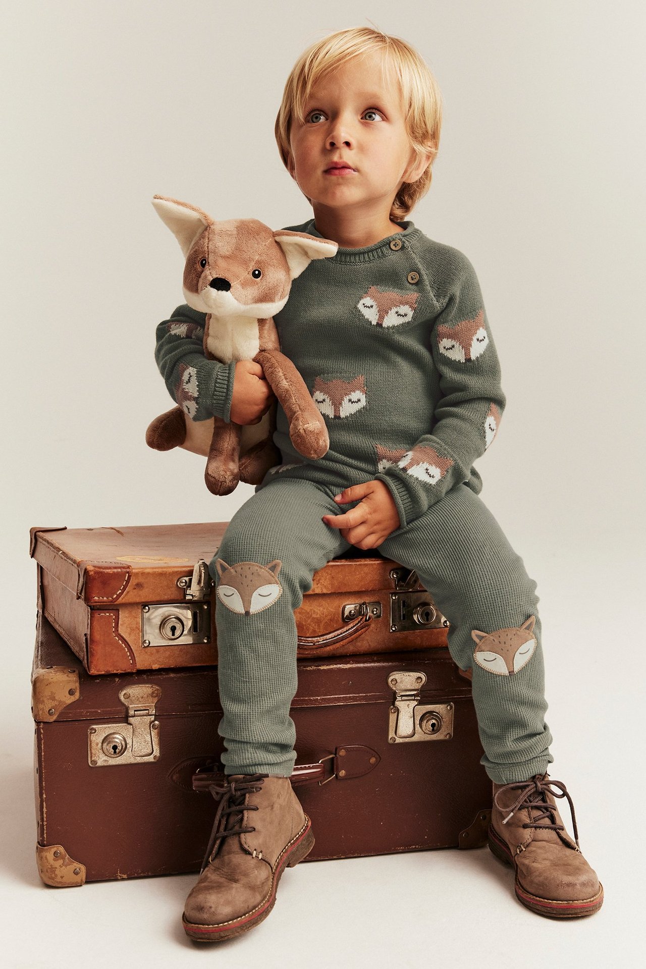 Vorderansicht: Kind in dunkelgrünen Waffel-Leggings mit Fuchs-Applikationen, passendem Pullover, braunen Stiefeln, hält Spielzeug.