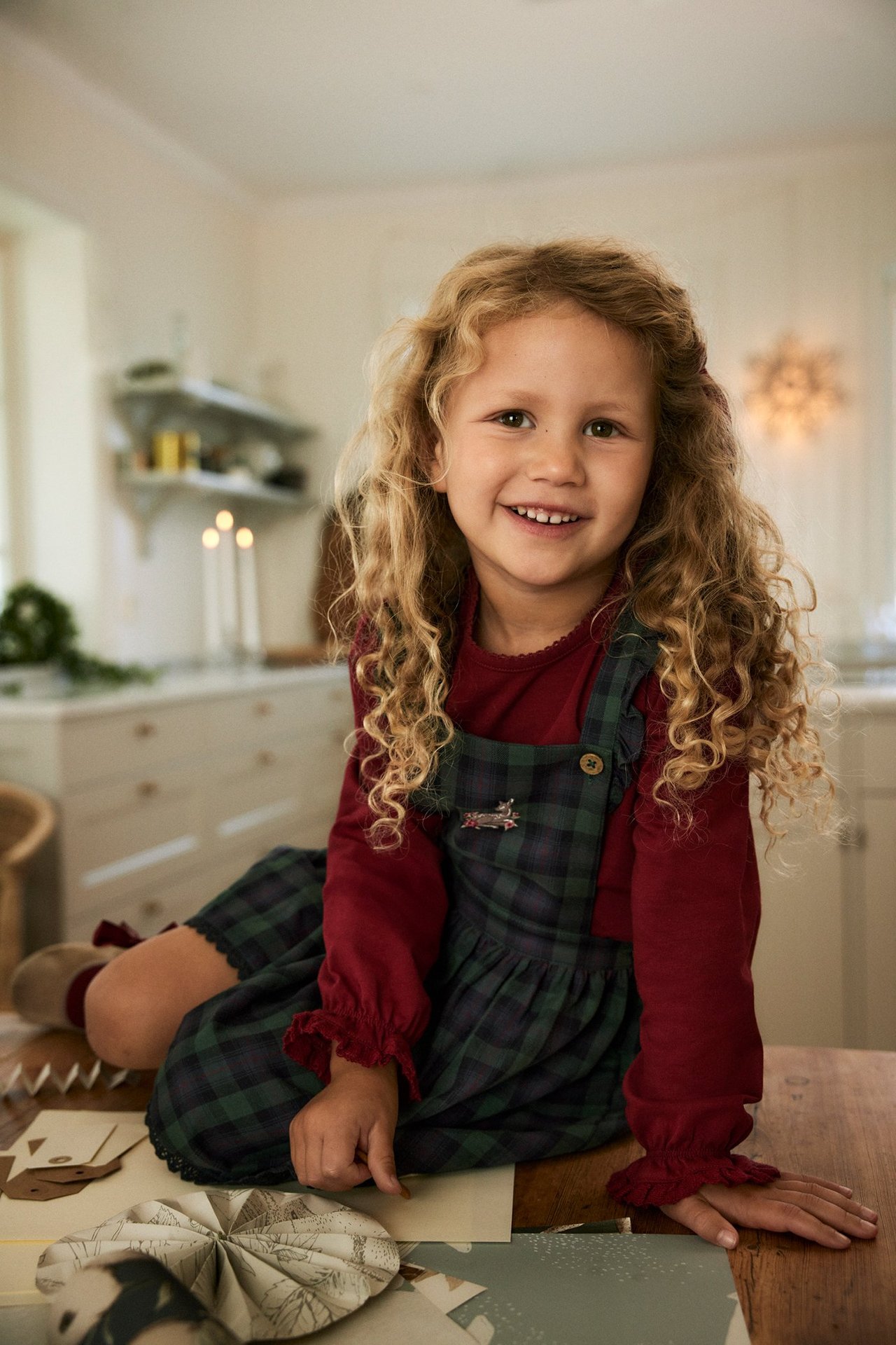 Smiling kid wears a blue plaid cotton dungaree dress over a red long-sleeved top, front view.