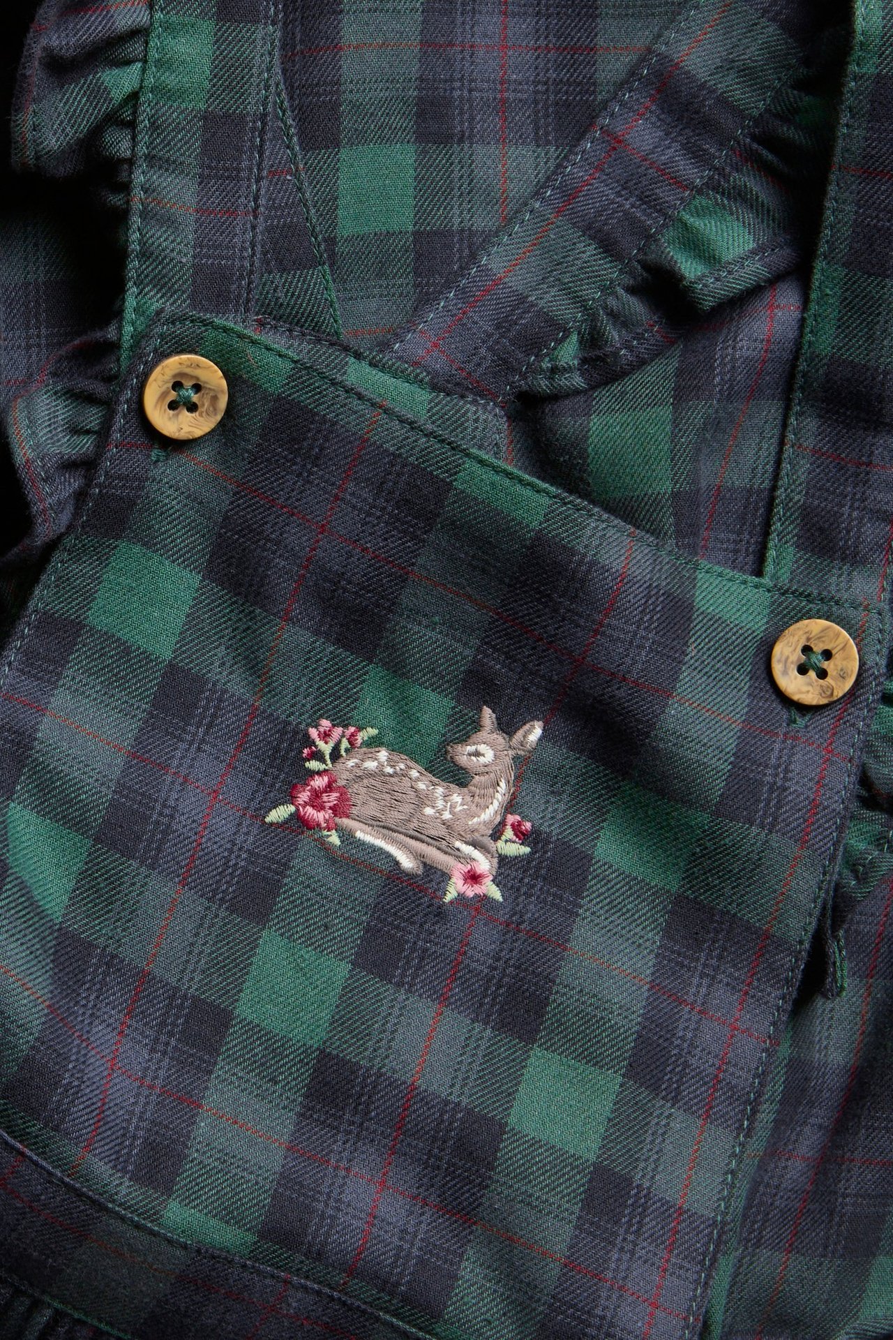 Close-up of a dark green and navy plaid suspender dress with fawn embroidery, wooden buttons.
