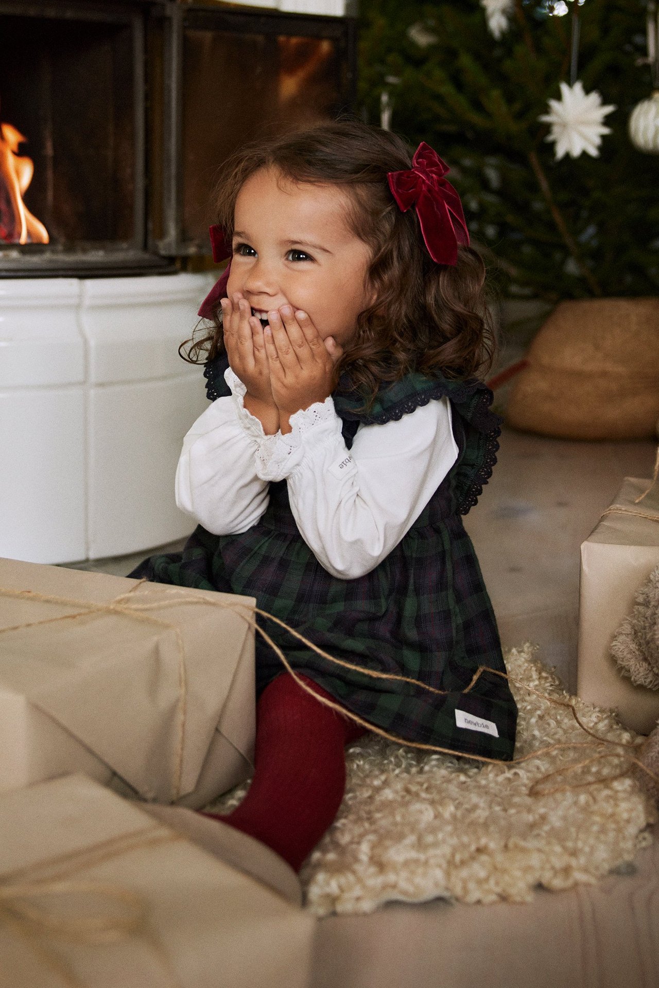 Front view, kid in dark green plaid pinafore dress, white long-sleeve top, red tights, red velvet bows.