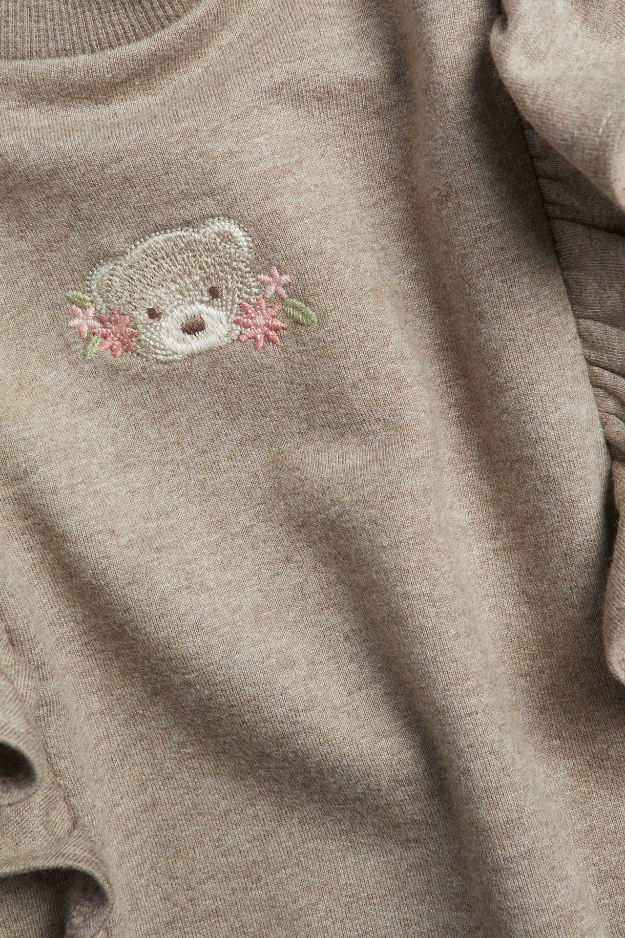 Nahaufnahme eines beigemelierten Kinder-Sweatshirts aus Baumwolle mit Rüschen und gesticktem Teddybär mit rosa Blumen.