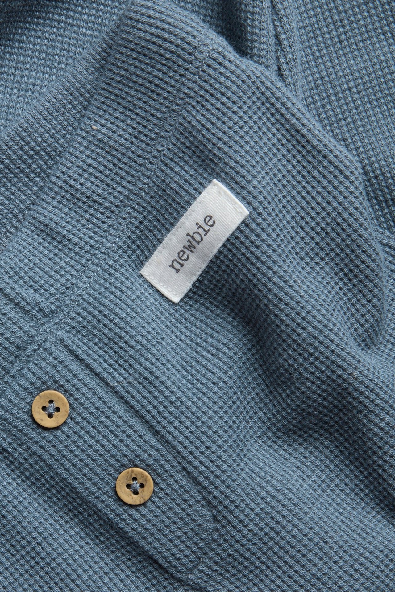 Nahaufnahme blauer Waffelstrick-Leggings mit dekorativen Holzknöpfen und weißem Newbie-Etikett.