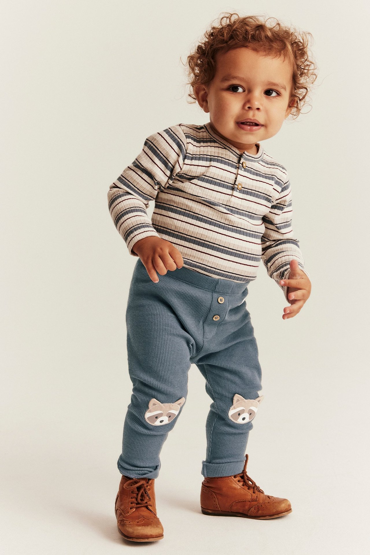 Vorderansicht eines Babys in blauen Waffelstrick-Leggings mit Waschbär-Knieapplikationen, gestreiftem Oberteil und braunen Stiefeln.