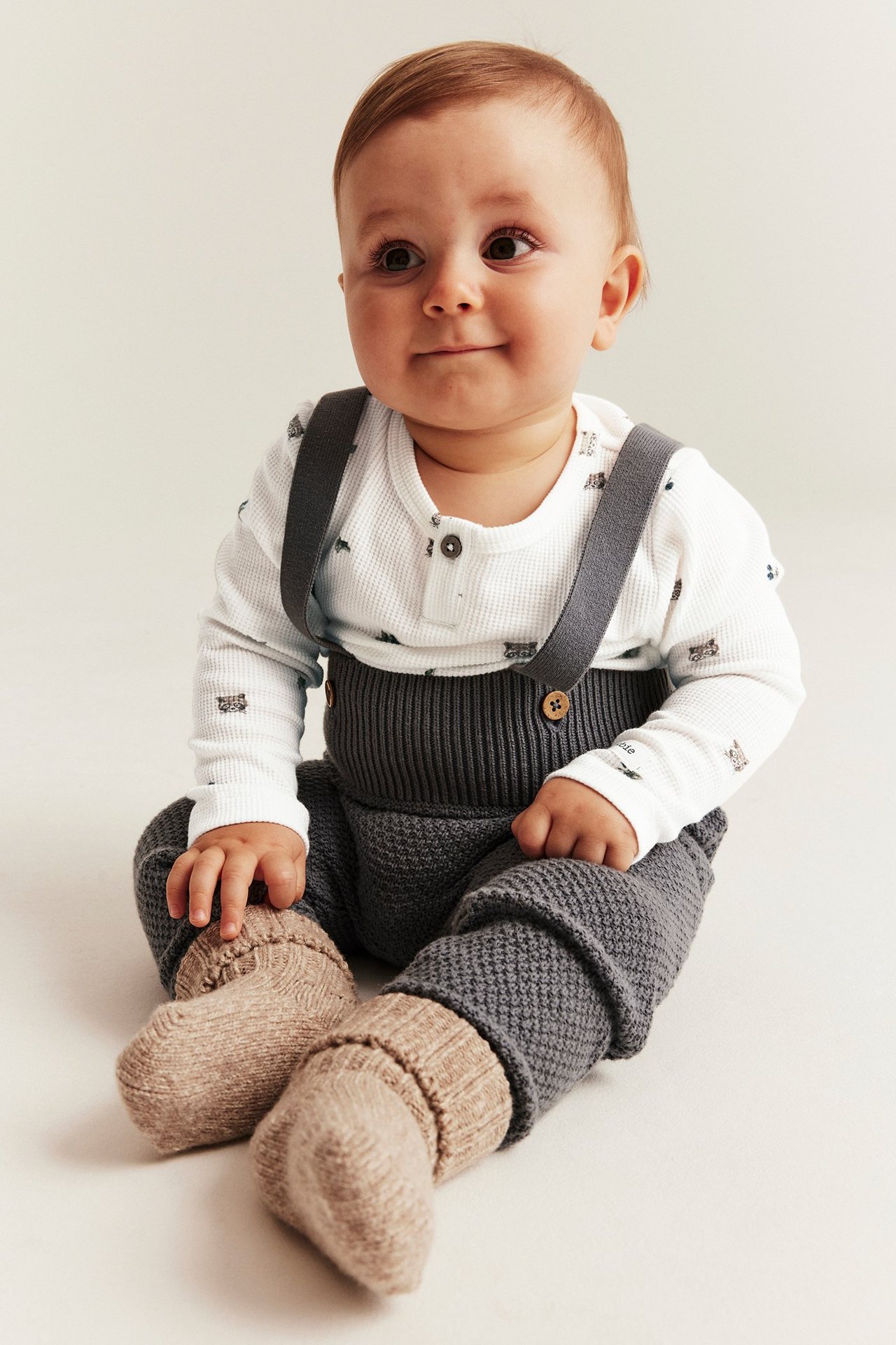 Baby trägt einen cremefarbenen Waffelstrick-Langarmbody mit Muster, dunkelgraue Strick-Latzhose und hellbraune Strickschühchen.