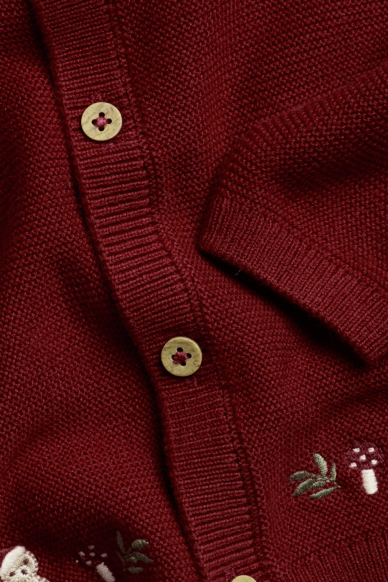 Close-up of a red knitted cotton cardigan with small white mushroom and green leaf embroidery, and light wooden buttons.