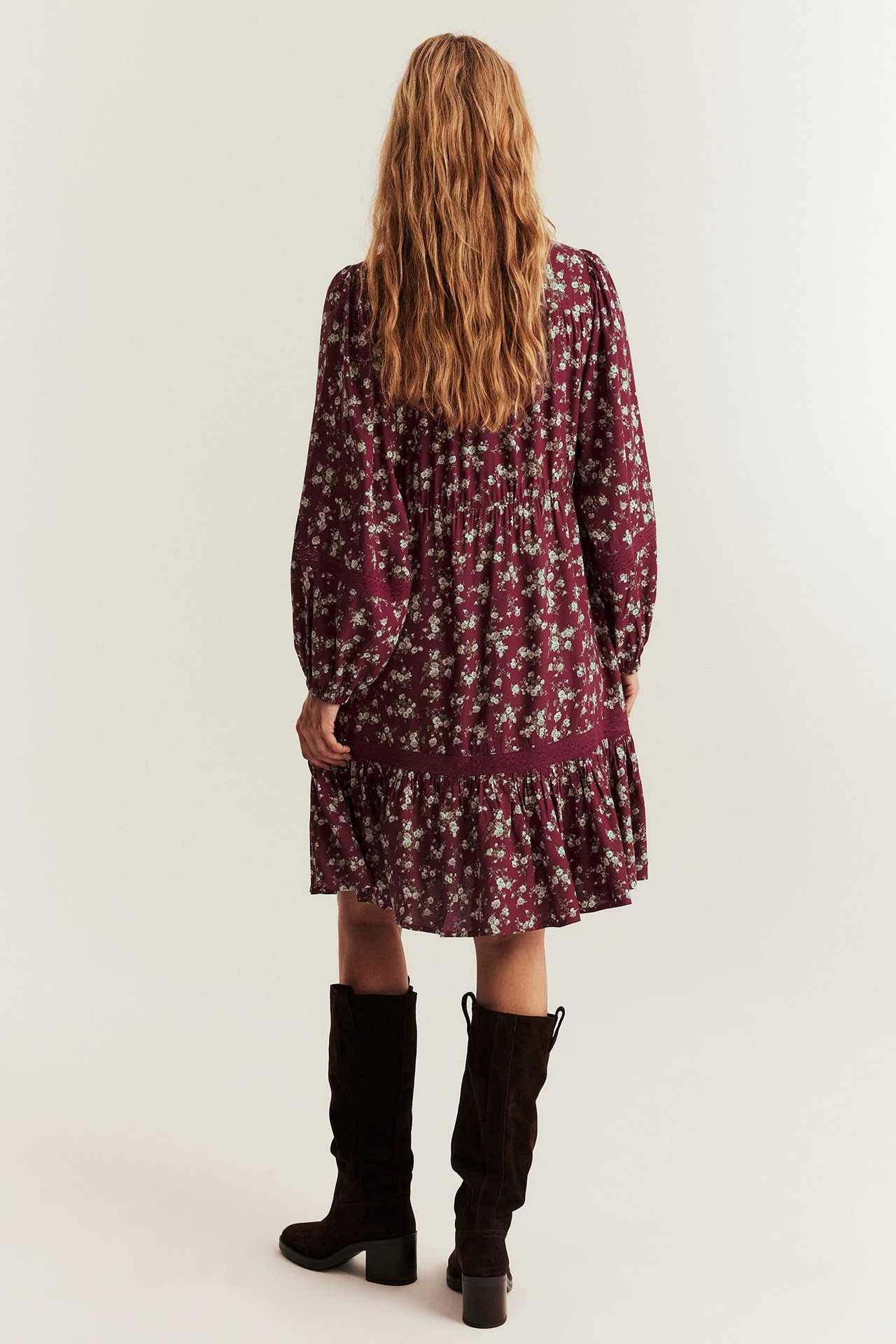 Back view: Woman in a dark red floral viscose knee-length dress with long sleeves and dark boots.