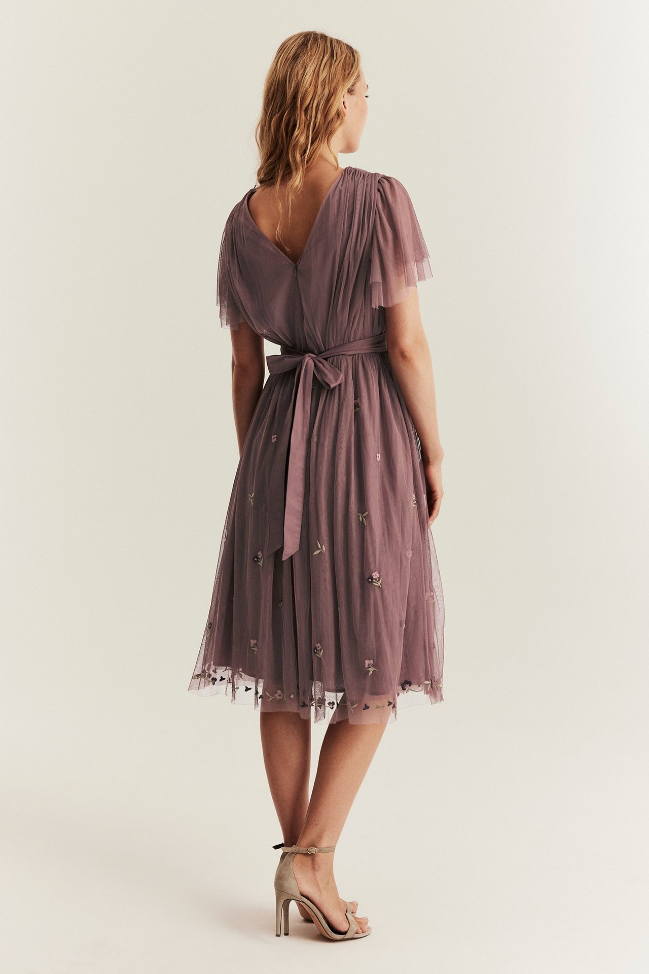 Rückenansicht, Frau in einem dunkelrosa bestickten Mesh-Midikleid mit Schmetterlingsärmeln und Rückenschnürung. Trägt beigefarbene Sandalen.