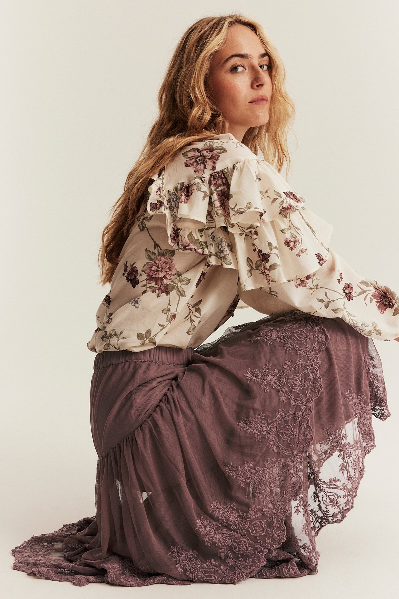 Seitenansicht einer knienden Frau in dunklem rosa besticktem Mesh-Rock und einer cremefarbenen floralen Rüschenbluse.