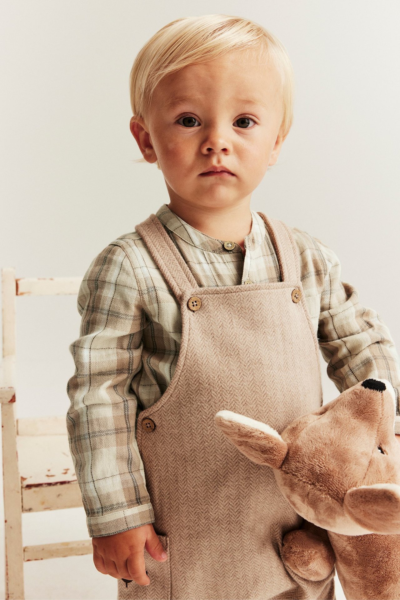 Vorderansicht eines Babys in einem beigen karierten Hemd-Body, beiger Fischgrät-Latzhose, hält ein braunes Stofftier.