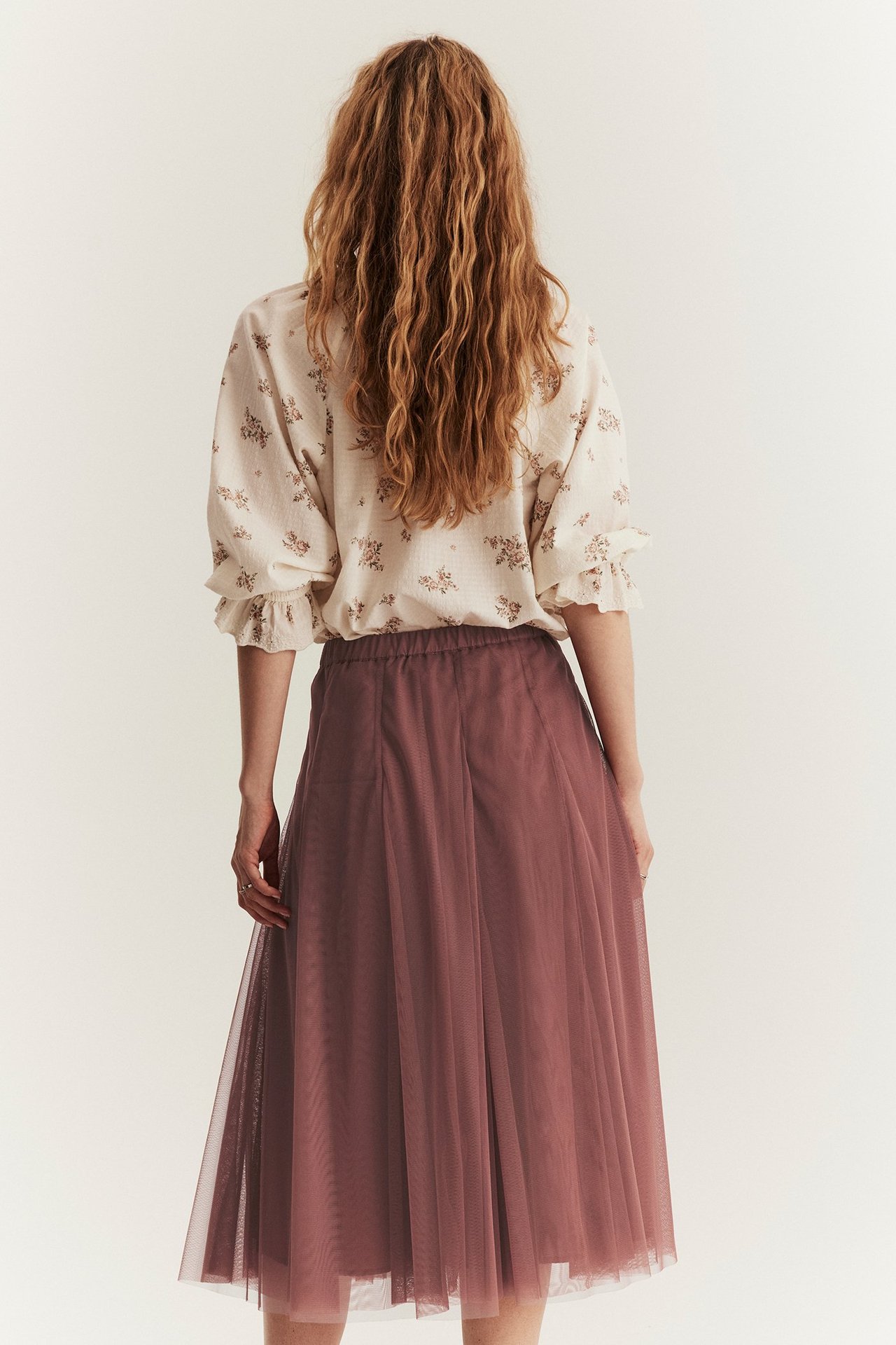 Rückenansicht einer Frau in einem dunkelrosa Mesh-Midirock mit Gummizug und einer hellen Blumenbluse.