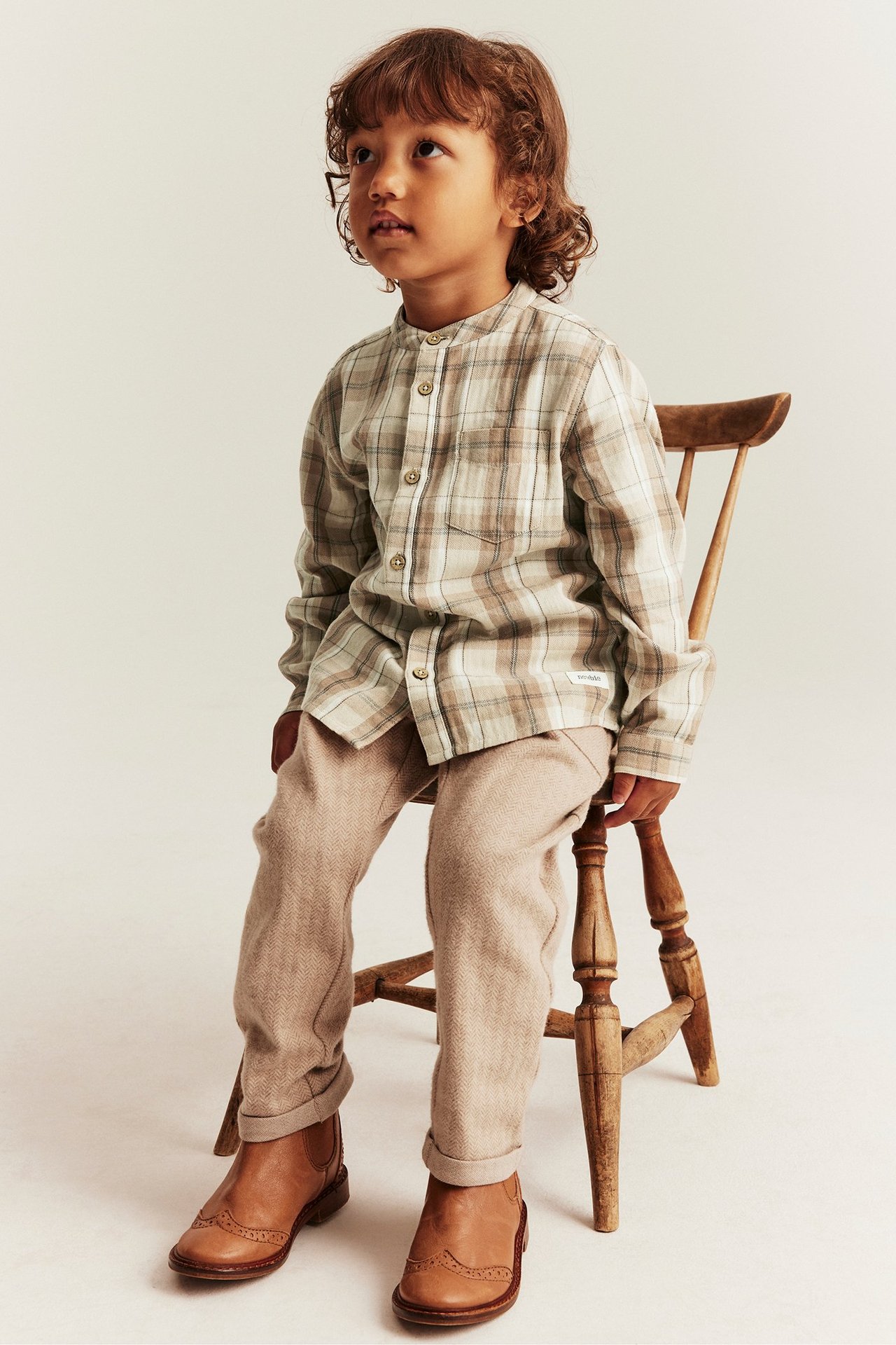 Front view of a kid in a beige plaid cotton shirt with mandarin collar, beige trousers, and brown boots.