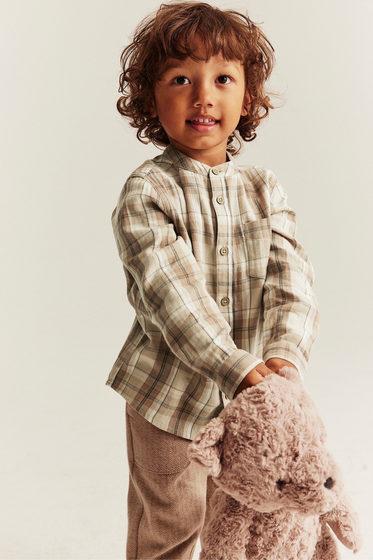Vorderansicht: Kind in beigem Karomuster-Baumwollhemd, Stehkragen, beige Hose, hält Teddybär.