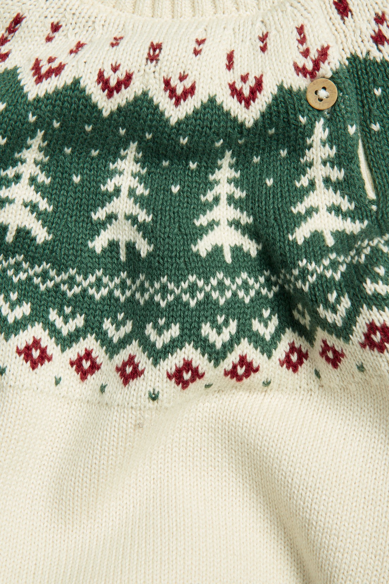 Nahaufnahme eines cremefarbenen gestrickten Weihnachtspullovers mit grünem Tannenbaum-Jacquardmuster und Holzknopf.