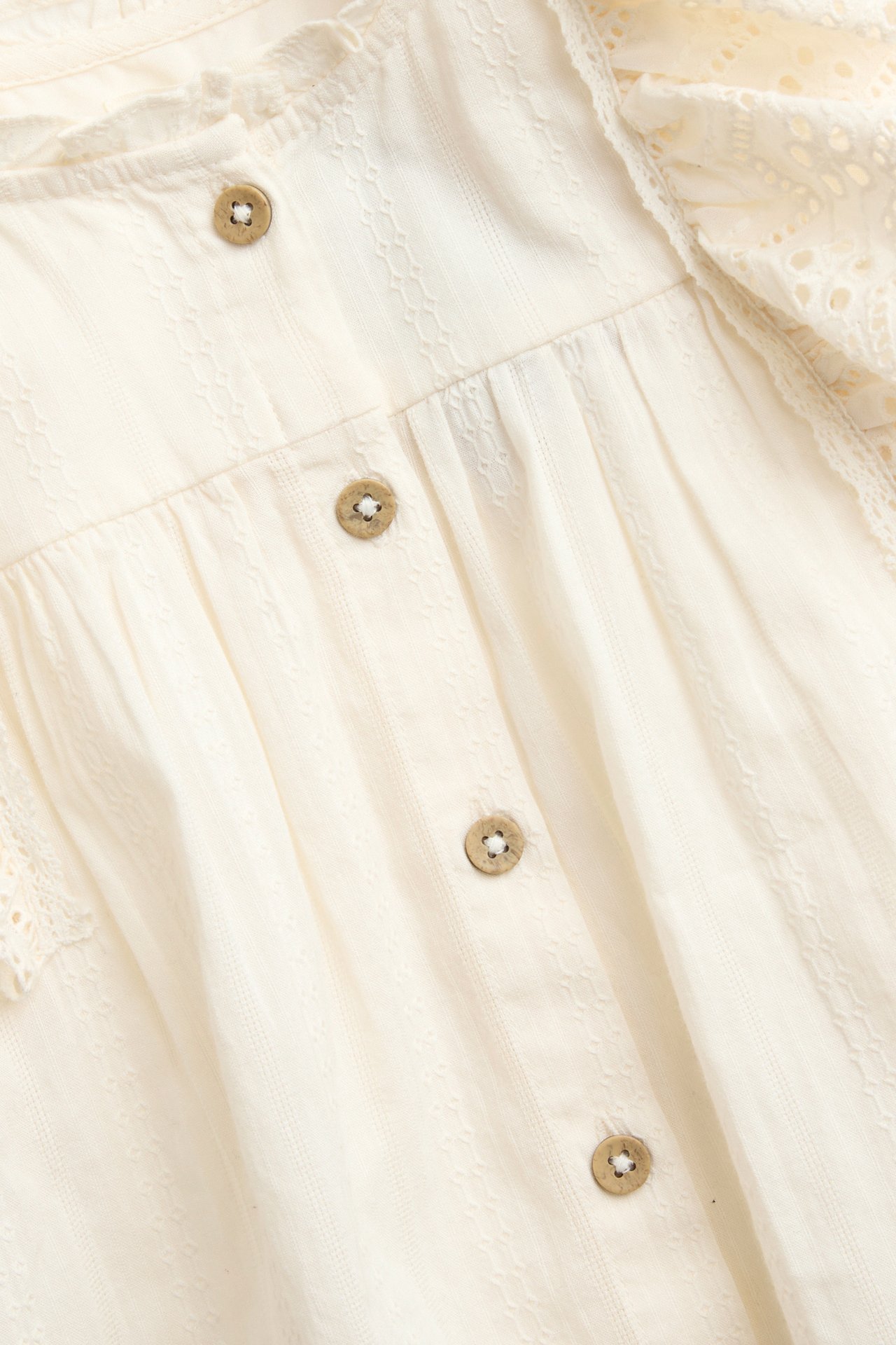 Close-up front view of an off-white cotton dobby blouse with embroidered ruffles and wooden buttons.