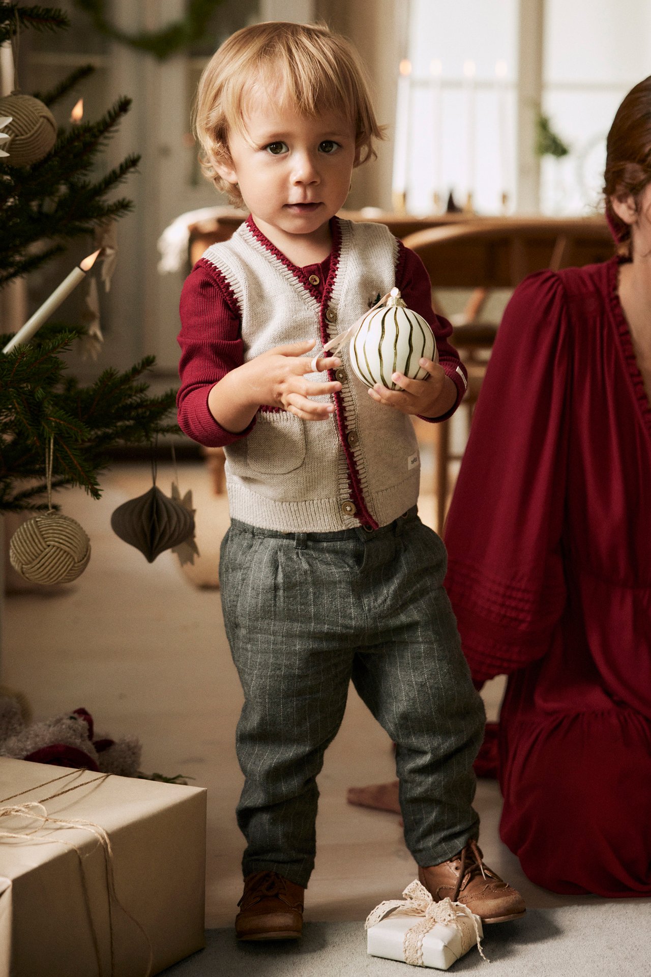 Front view: Kid wears beige melange knitted vest over red shirt, grey plaid trousers, brown shoes, holding ornament.
