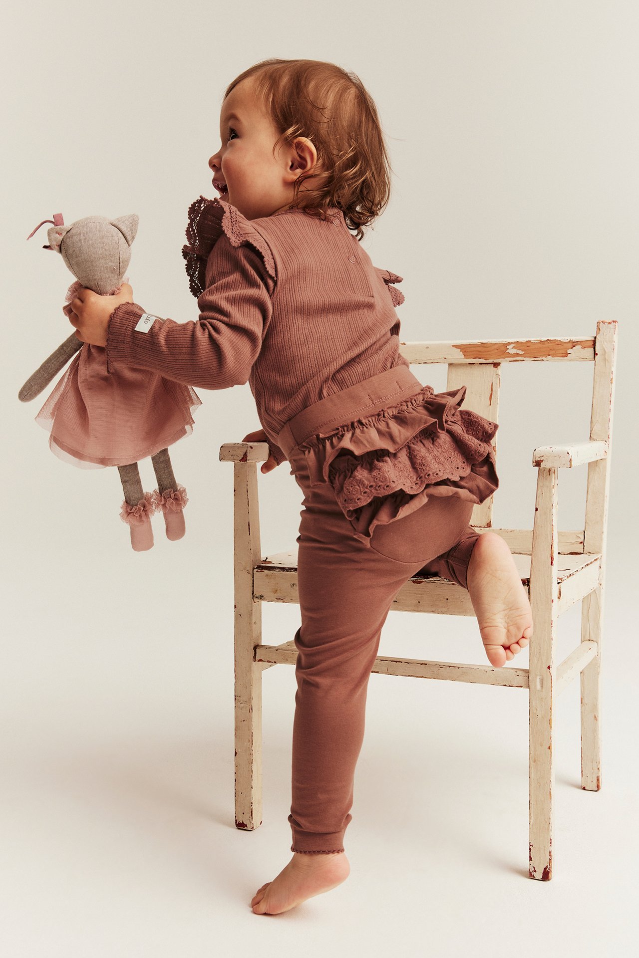 Side view of a baby in pink leggings with back ruffles and lace, matching top, holding a doll.