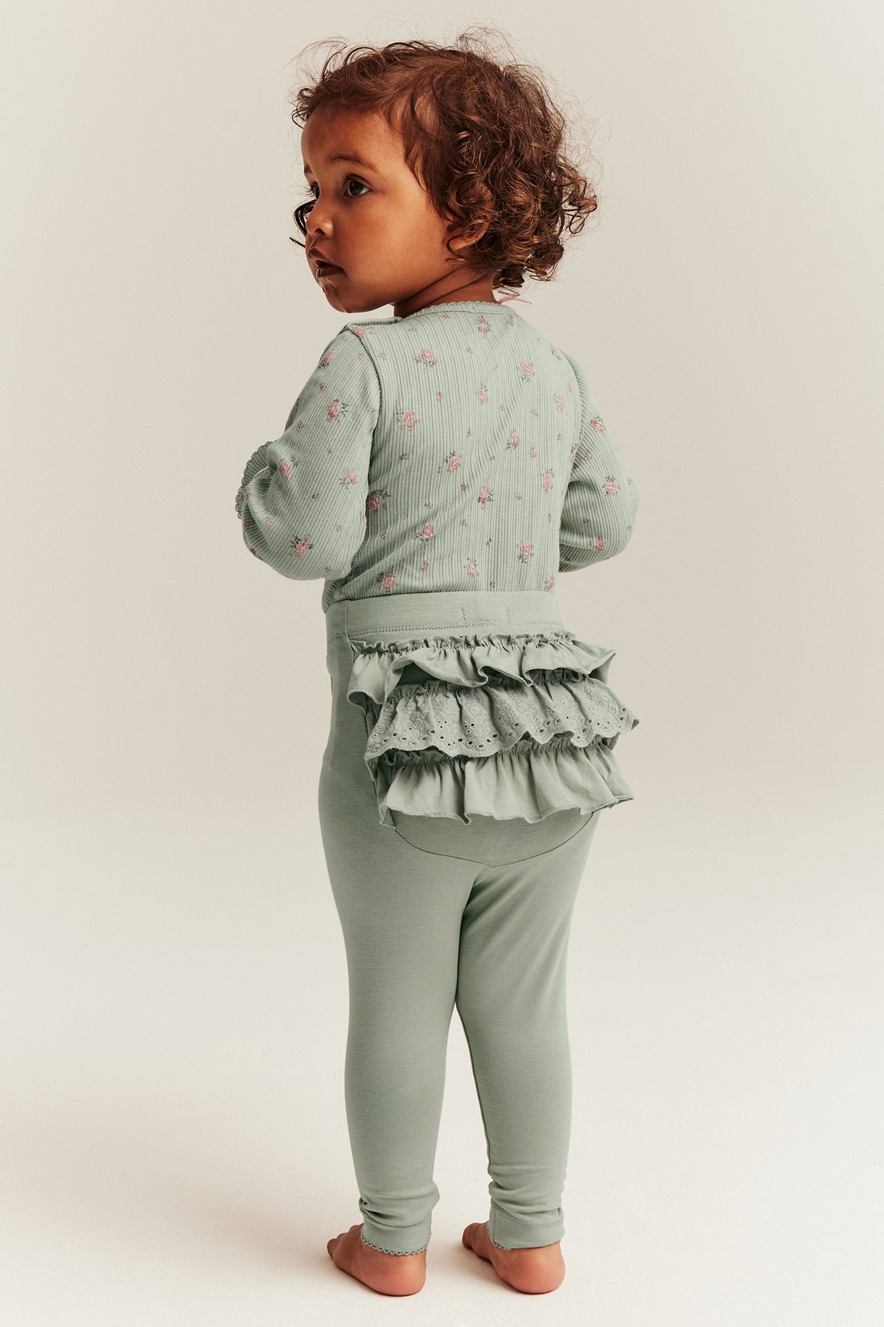 Rückenansicht eines Babys in grünen Rüschen-Leggings mit Spitze und einem geblümten Langarmshirt.