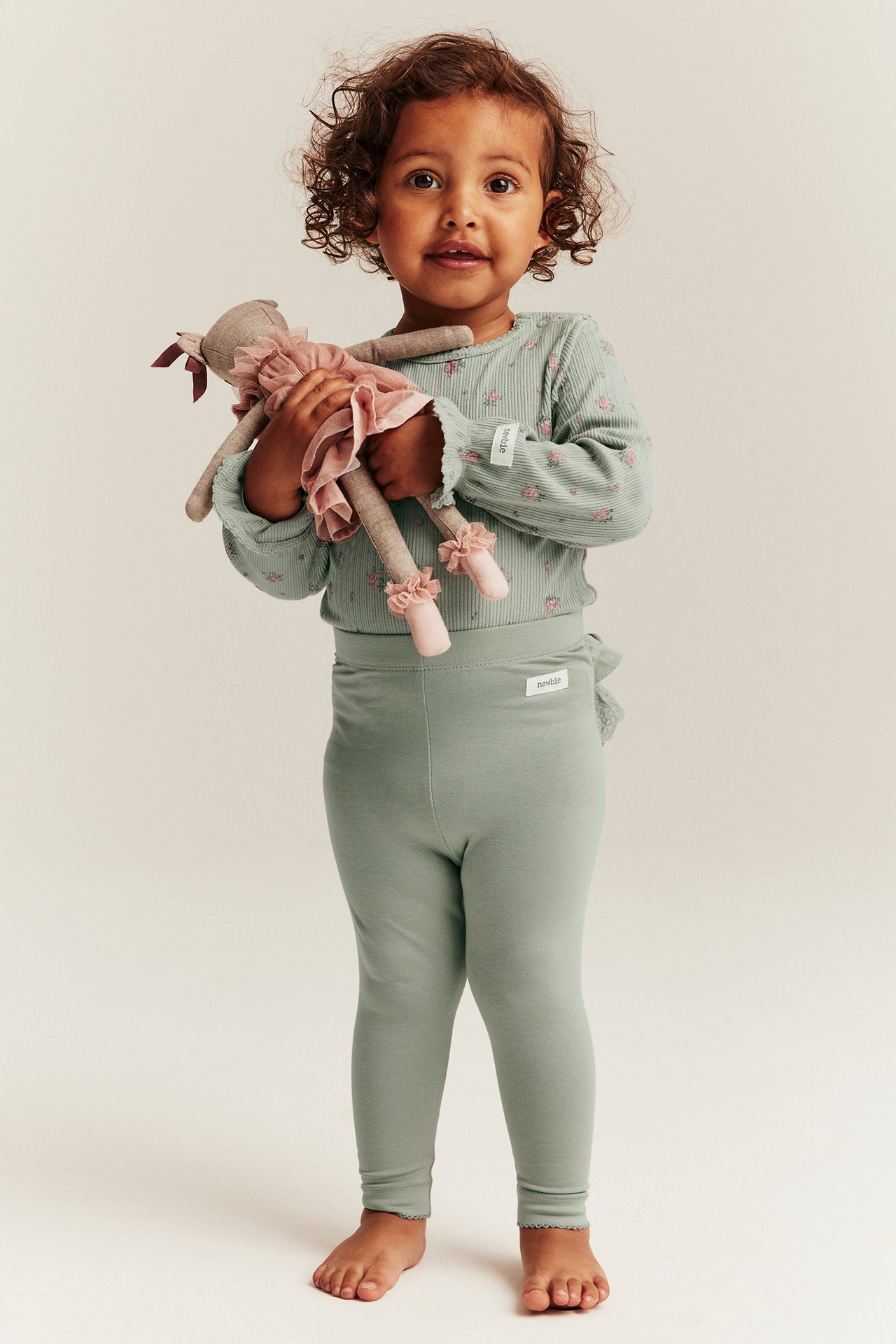 Vorderansicht eines Kindes in grünen Leggings mit Rüschen und Spitze hinten, und einem hellgrünen geblümten Top, hält eine Puppe.