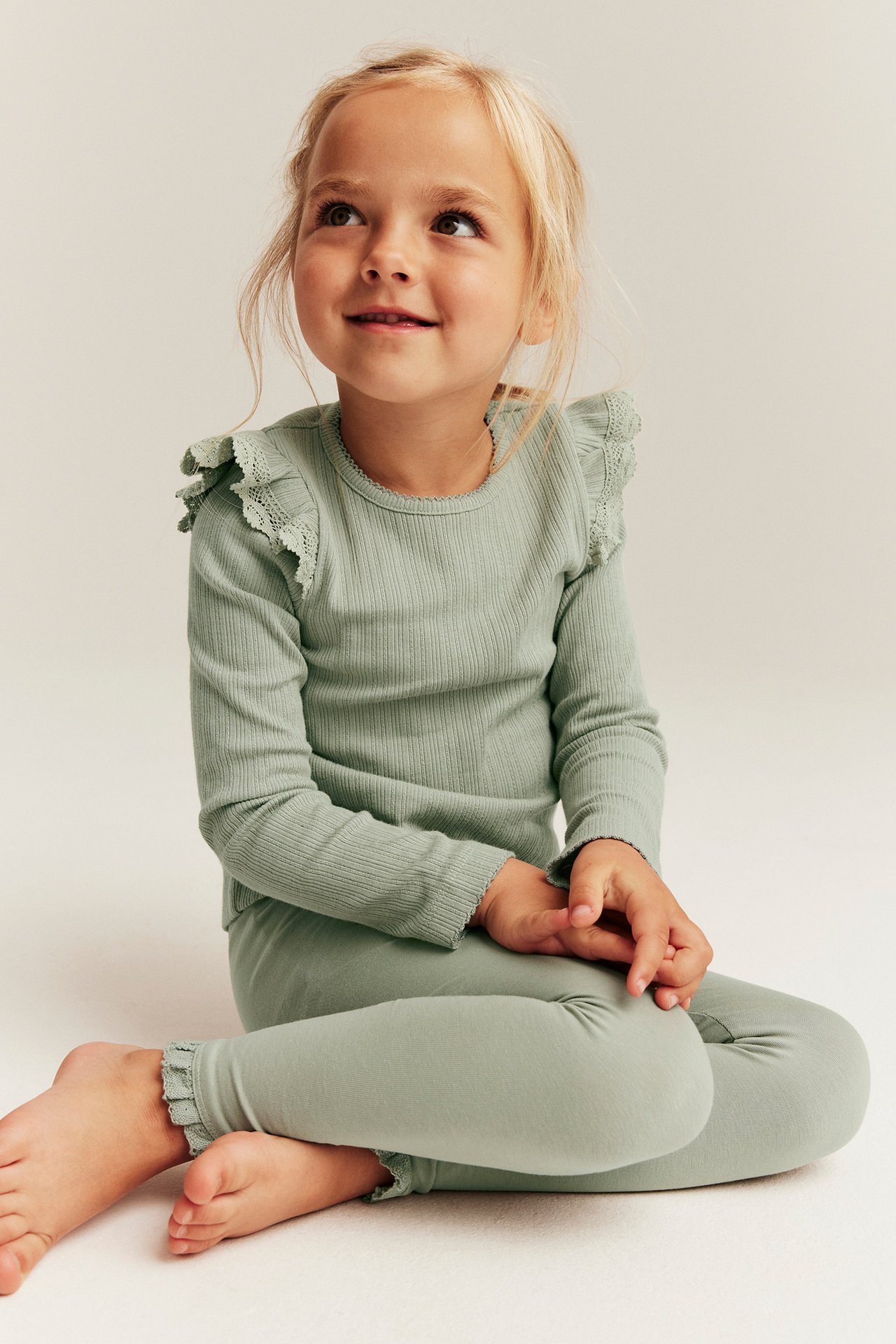 Front view of a sitting kid wearing green leggings with lace ruffles and a matching ribbed long-sleeve top.