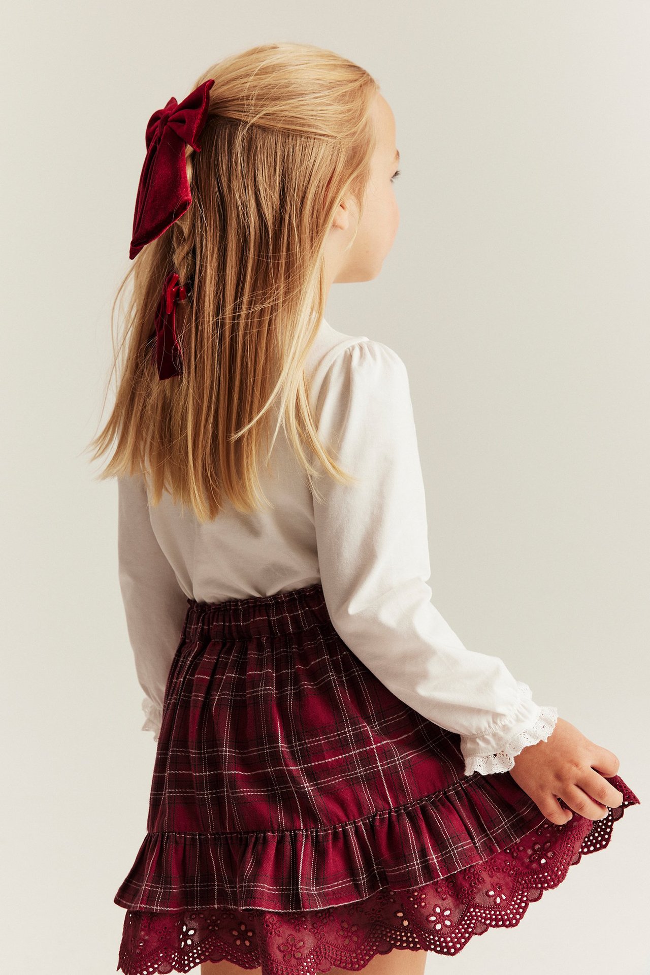 Girl in profile, red plaid skirt with ruffles, white blouse, and large red hair bow.