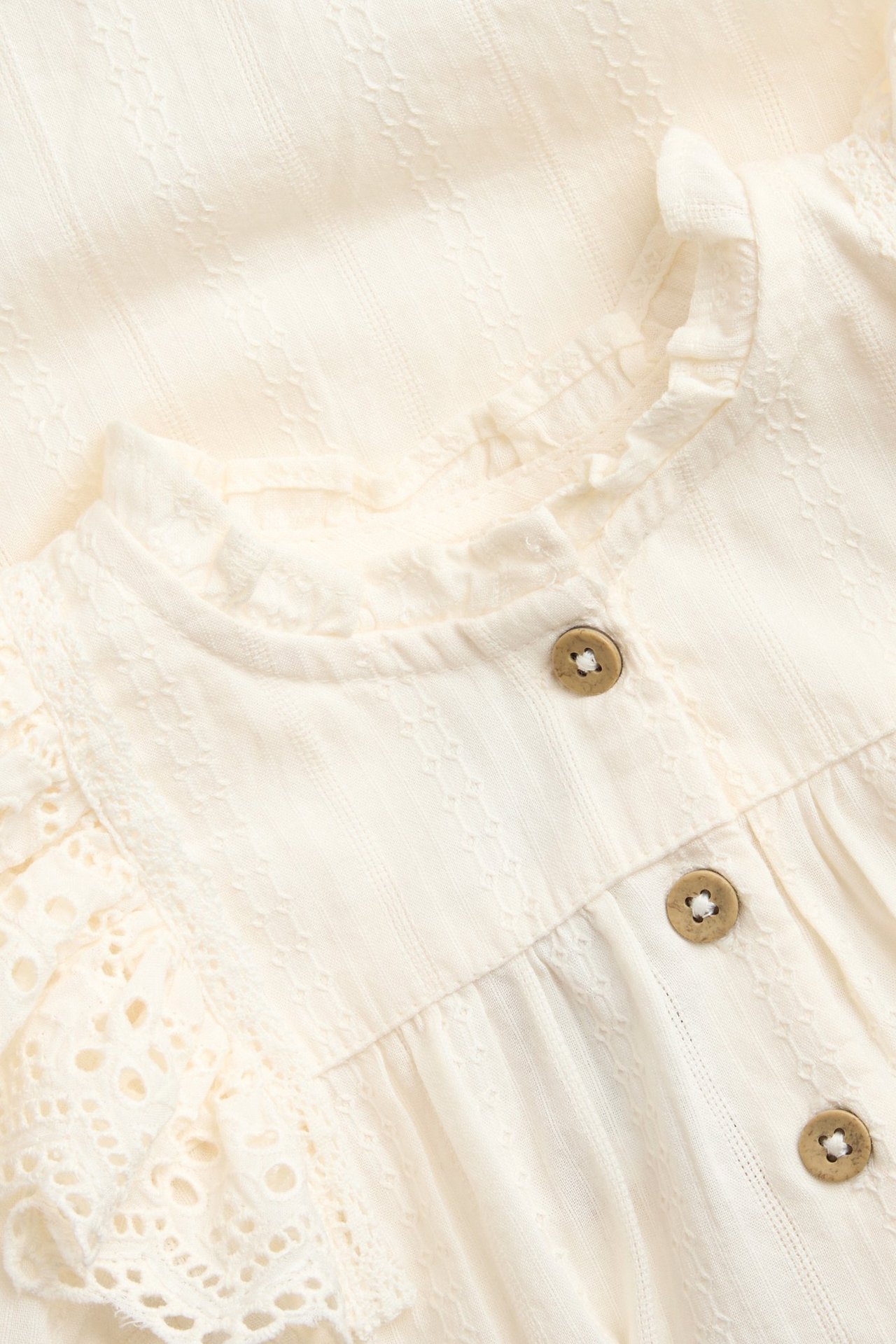 Nahaufnahme, Vorderansicht: Off-white Baby-Baumwolldobby-Body mit bestickten Rüschen und Vorderknöpfen.