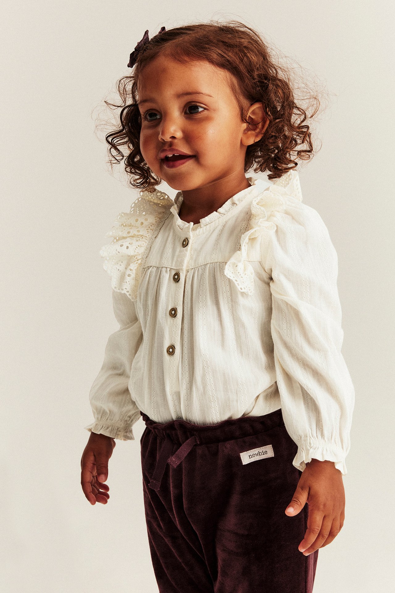 Front view of a kid wearing an off-white cotton bodysuit with embroidered ruffles and dark purple velvet trousers.