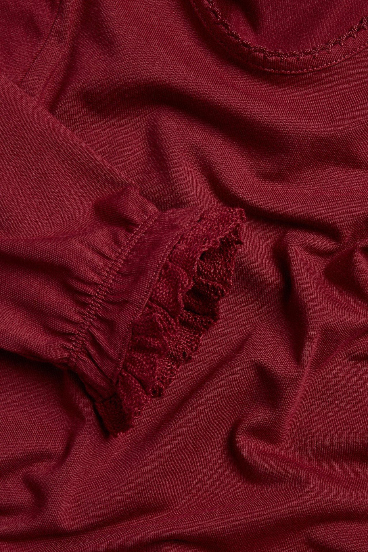 Close-up of a red long-sleeved baby bodysuit with picot neckline and lace-trimmed ruffled cuffs.