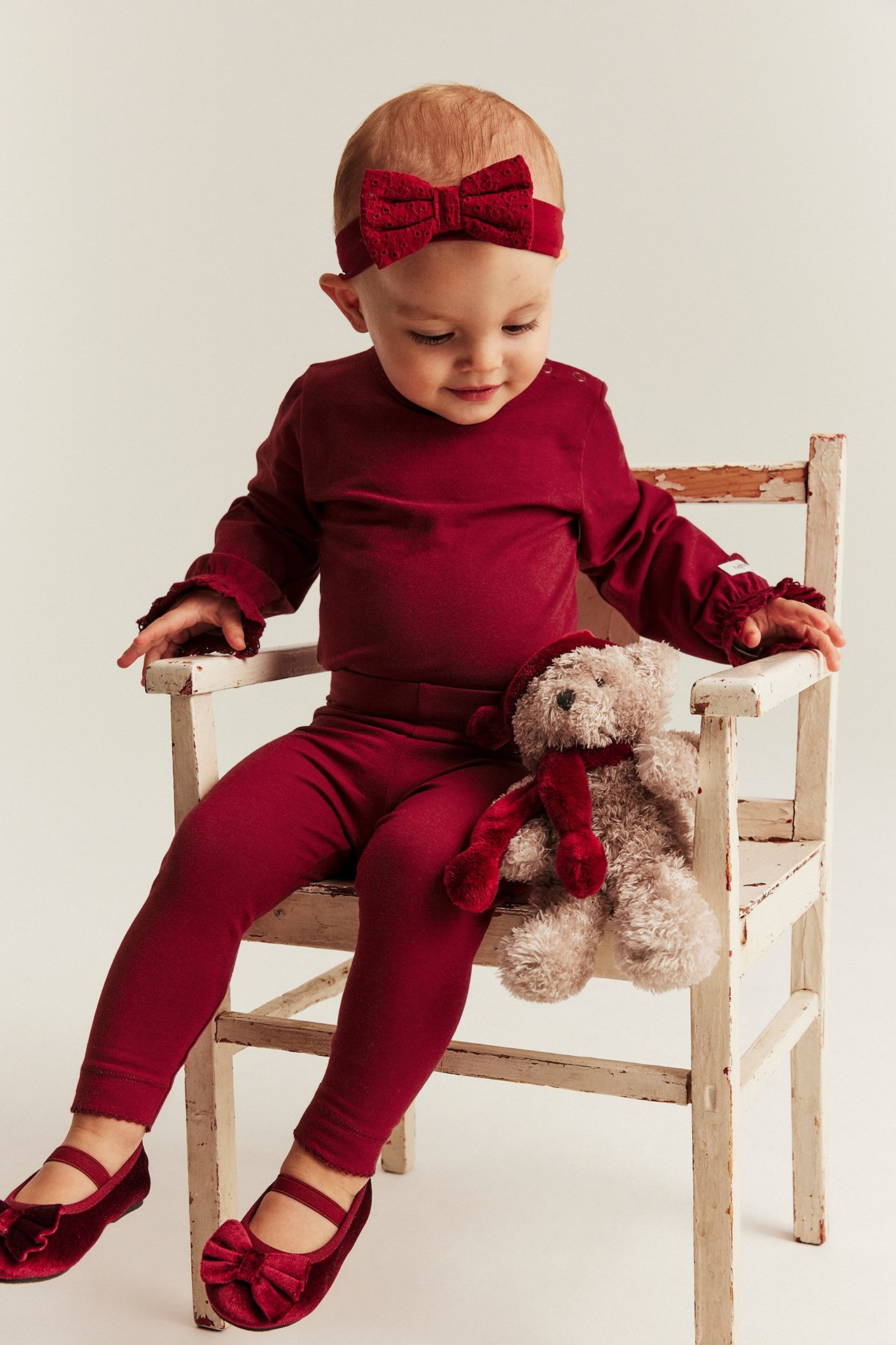 Baby girl in red long-sleeve body with bow headband, sitting on a white wooden chair.