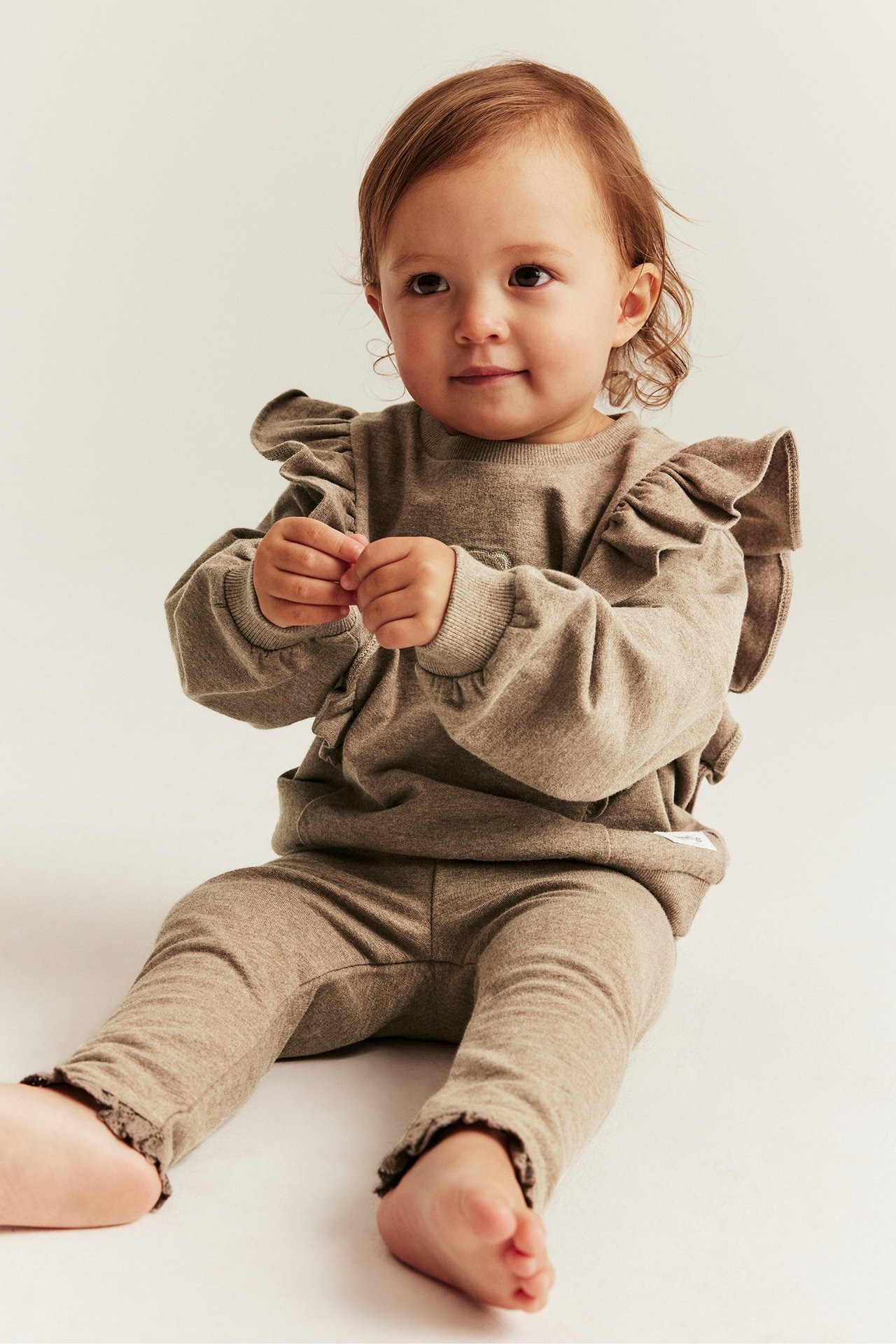 Front view: Baby in brown melange leggings with lace trim and a matching ruffled long-sleeved top.