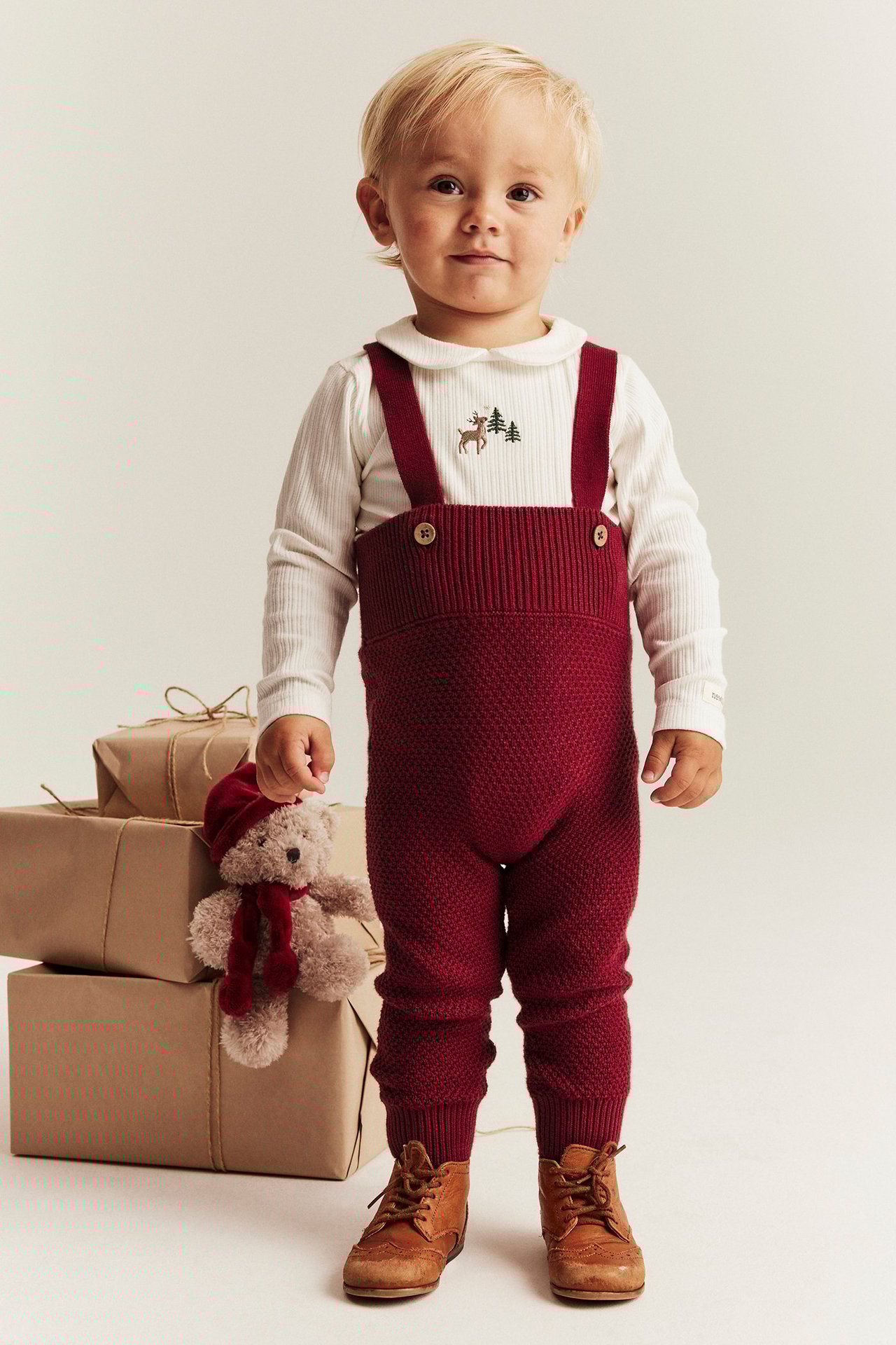 Girl in white sweater with reindeer and fir, red ribbed knitted overalls with adjustable straps.