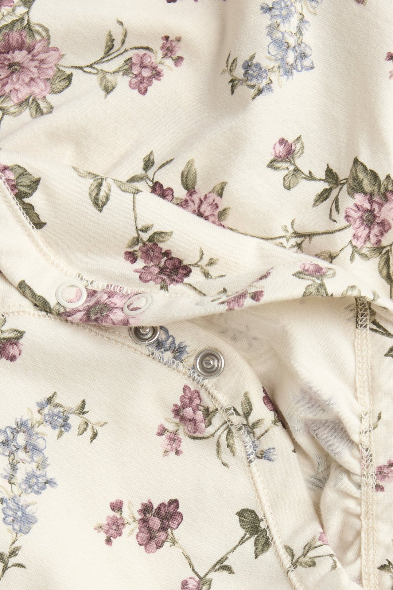 Nahaufnahme eines hellbeigen Baby-Bodys mit lila-blauem Blumenmuster und Druckknöpfen.