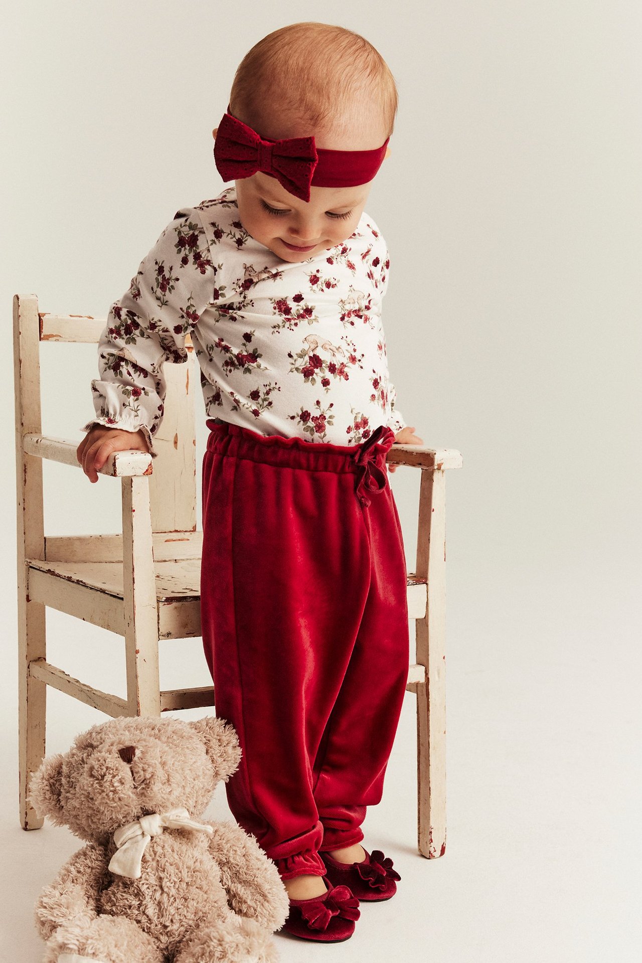 Child in floral cotton bodysuit with ruffle, red headband, velvet pants, shoes, with teddy bear.