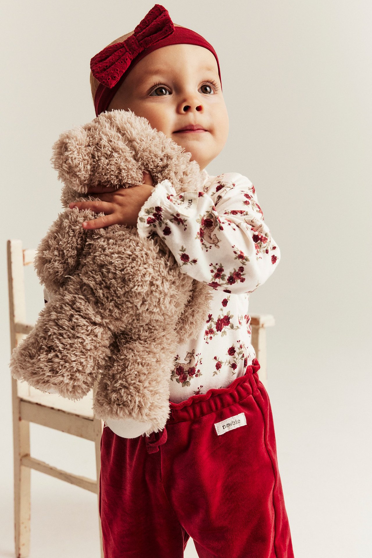 Child in offwhite floral body with ruffle, red pants, holding teddy bear, front view.
