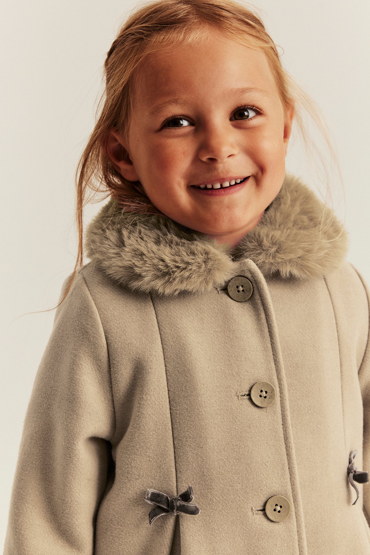 Close-up front view of a kid wearing a beige coat with a faux fur collar and three buttons.