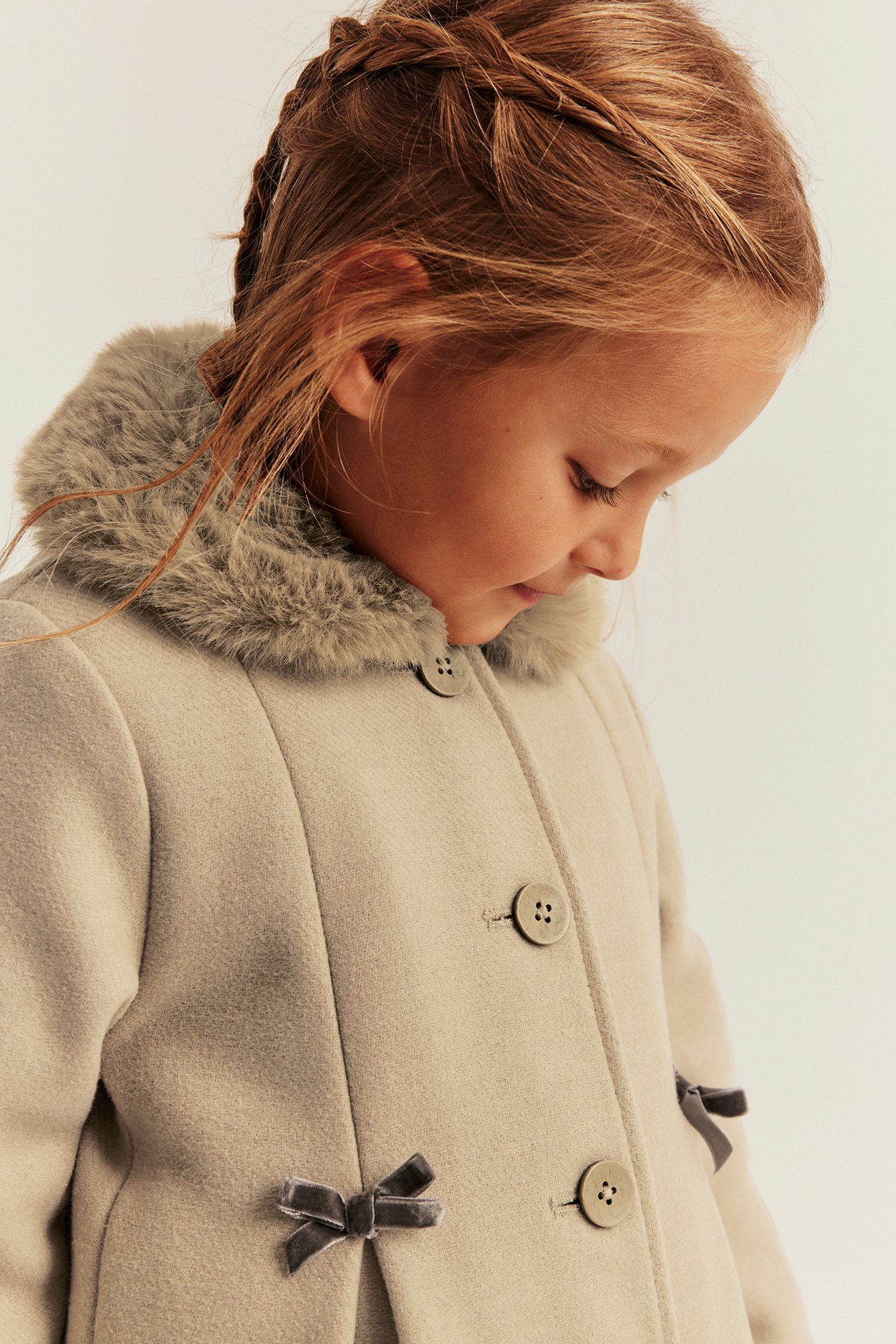 Close-up front view of a kid wearing a light beige coat with a faux fur collar and velvet bows.