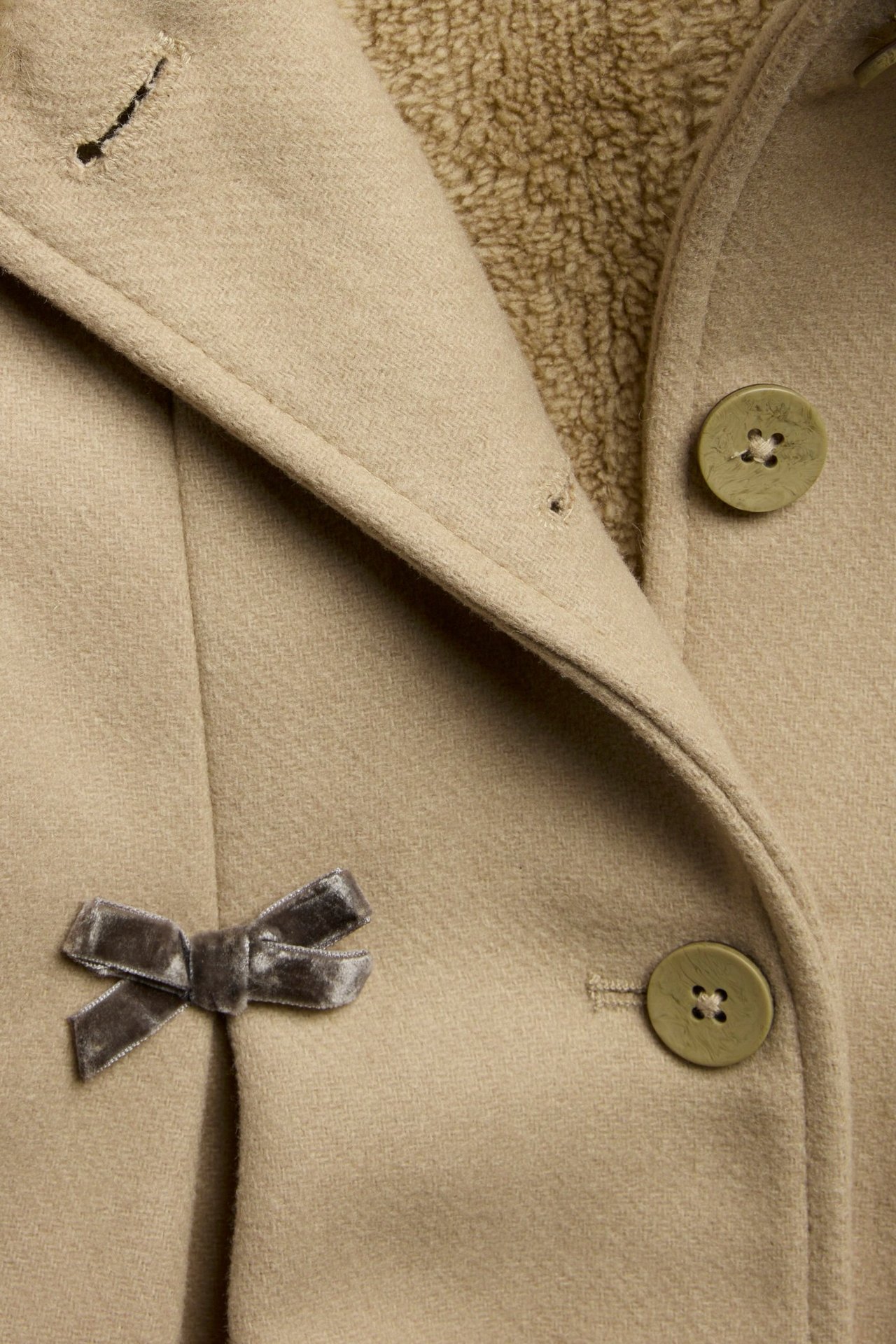 Nahaufnahme Vorderansicht eines beigen Wollmix-Mantels mit Teddyfutter, grünen Knöpfen und grauer Samtschleife.