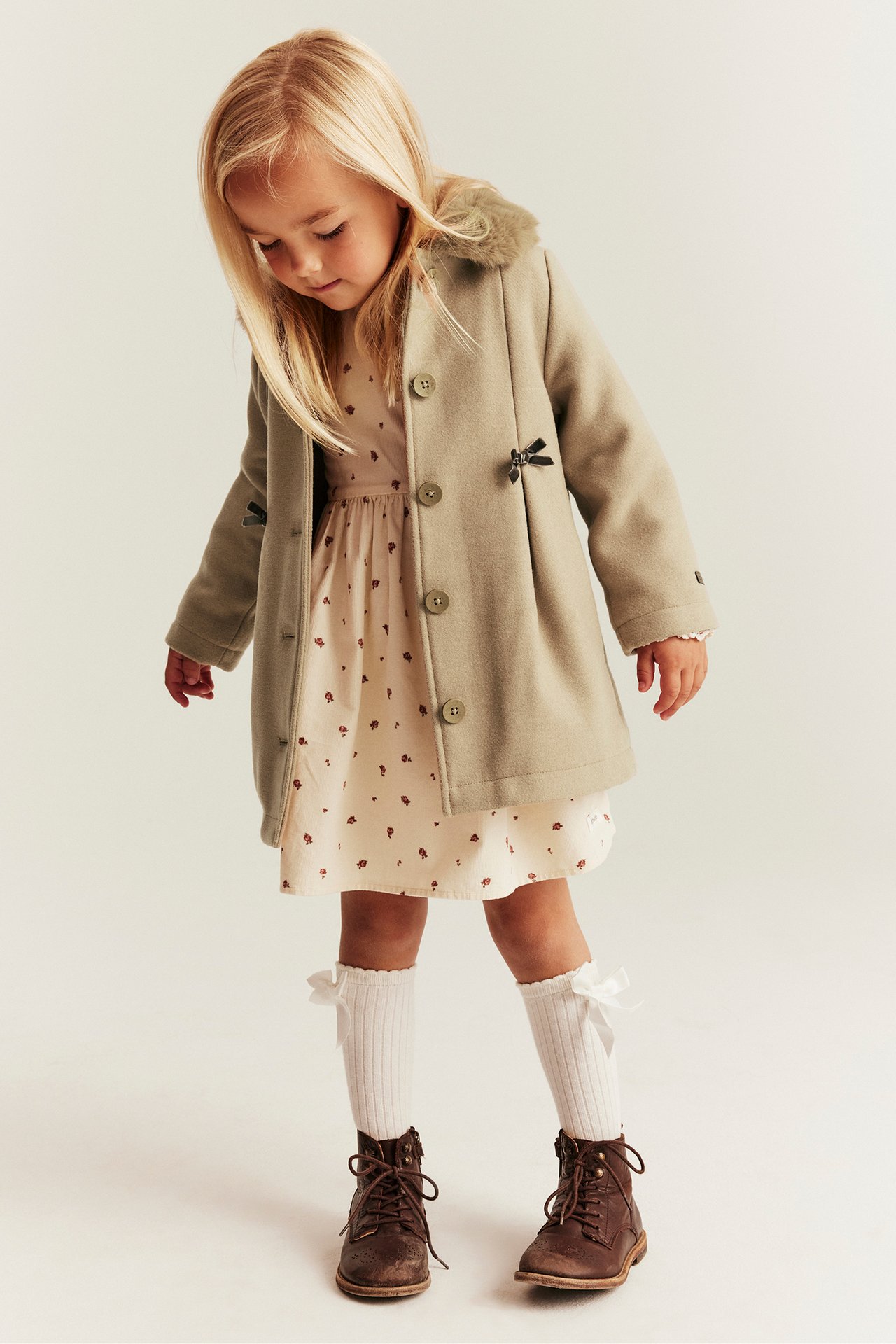 Vorderansicht: Kind in beigem Mantel mit Schleifen, cremefarbenem bedrucktem Kleid, weißen Schleifensocken und braunen Schnürstiefeln.