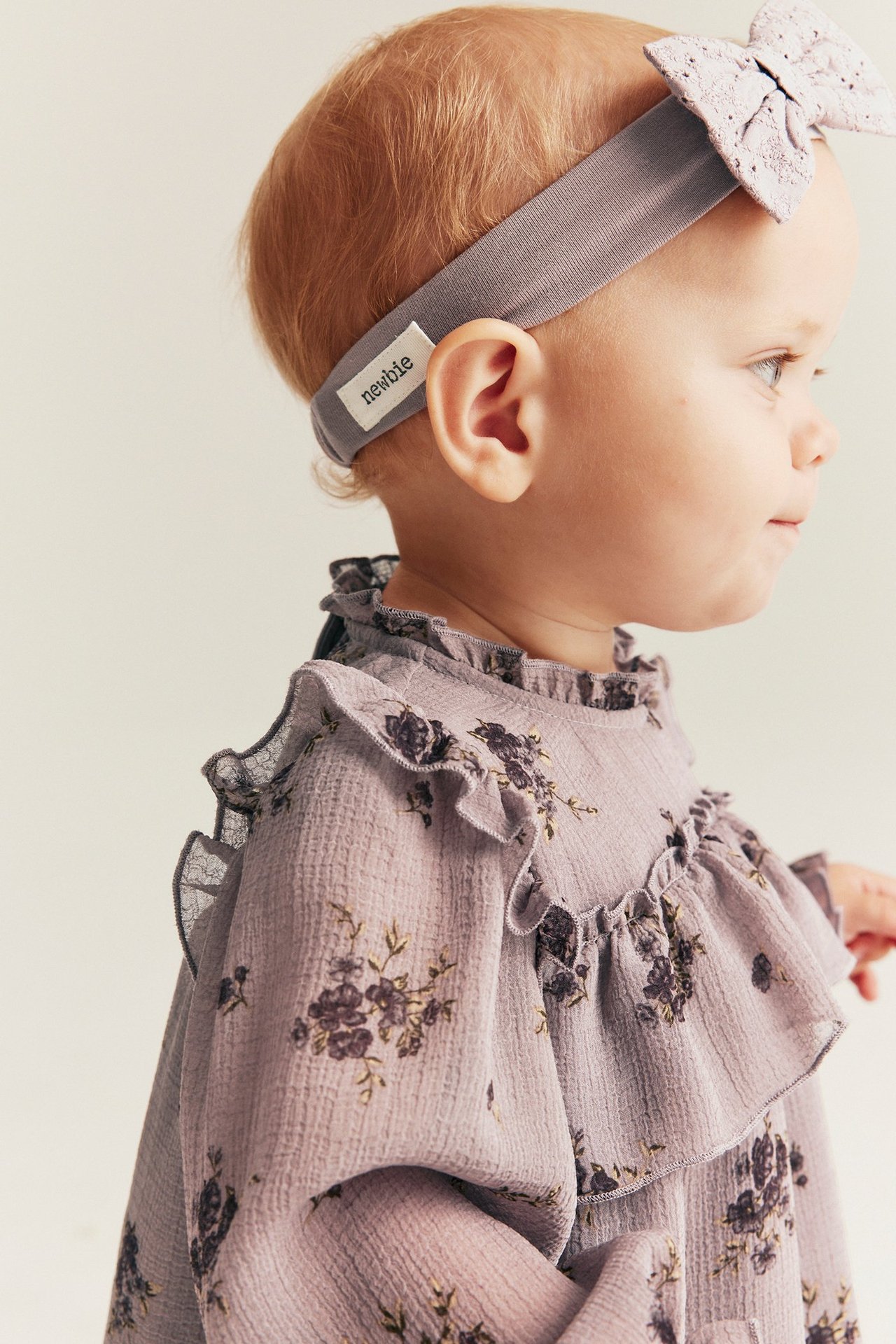 Nahaufnahme, Seitenansicht eines Babys, das ein helllila Jersey-Haarband mit Spitzenschleife und ein geblümtes Rüschenoberteil trägt.