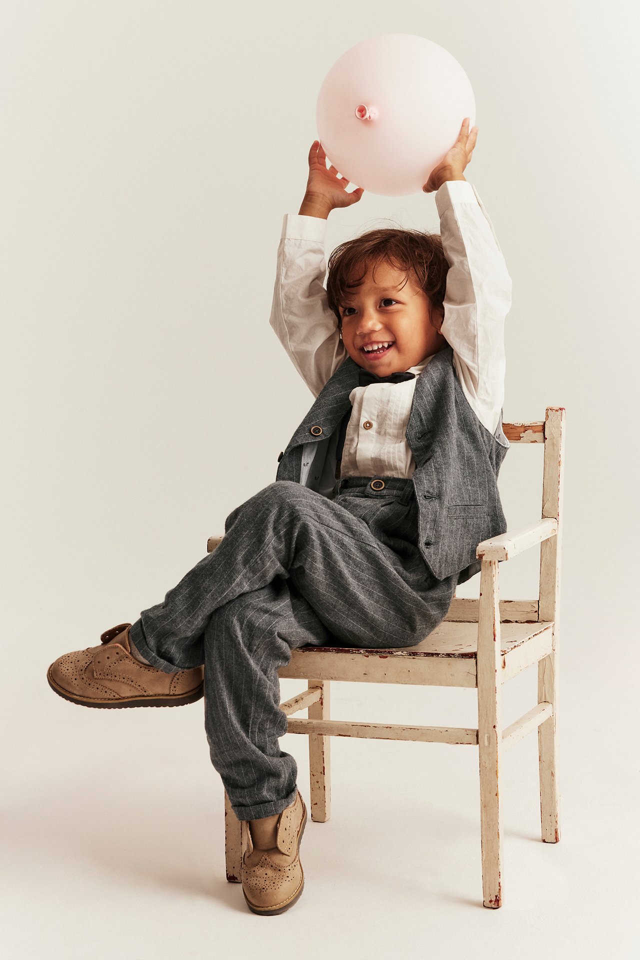 Kid wears a grey pinstripe woven vest, white shirt, matching trousers, tan shoes, holding a pink balloon.