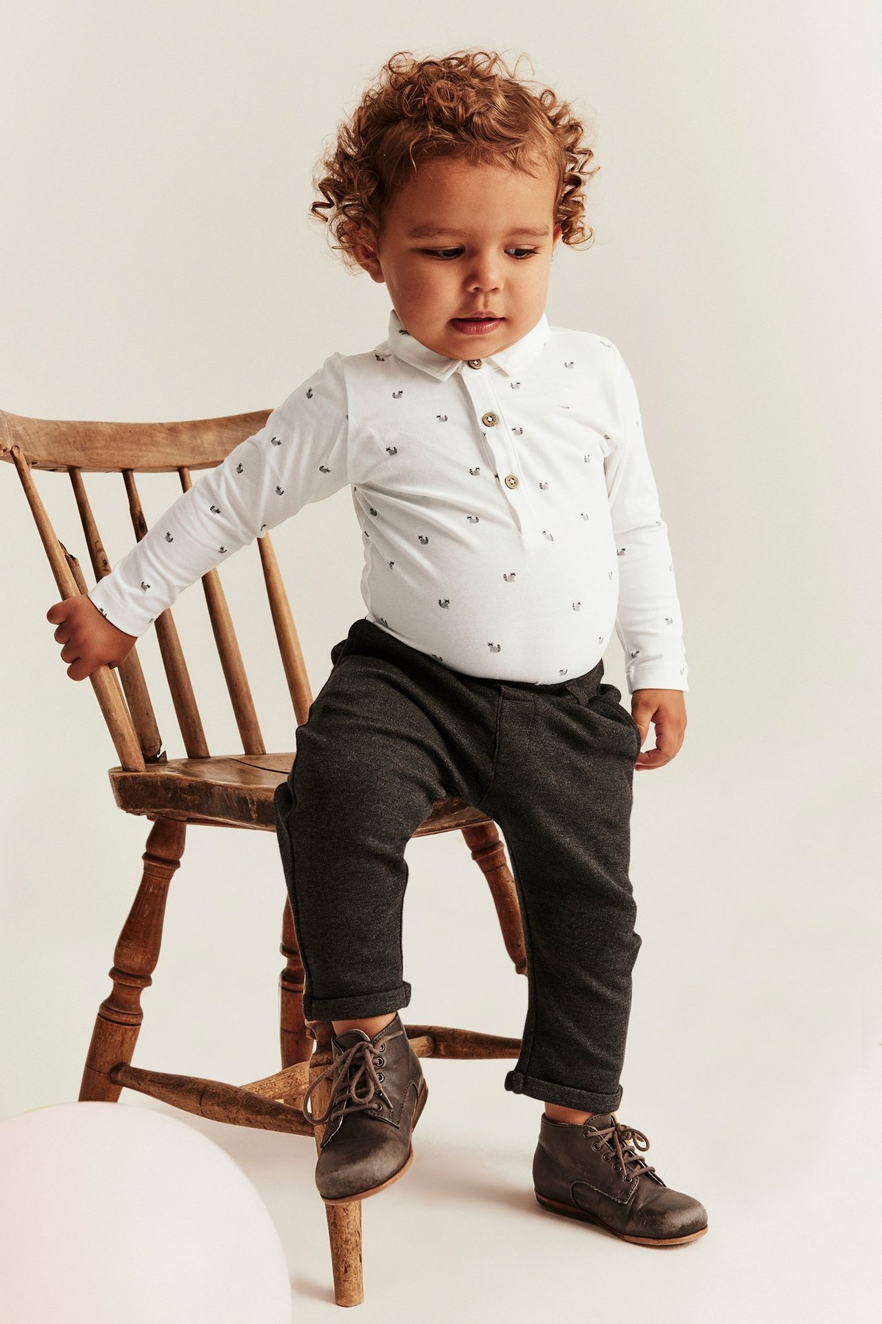 Front view of a baby wearing an offwhite long-sleeved patterned bodysuit with a collar, dark grey trousers, and brown boots.