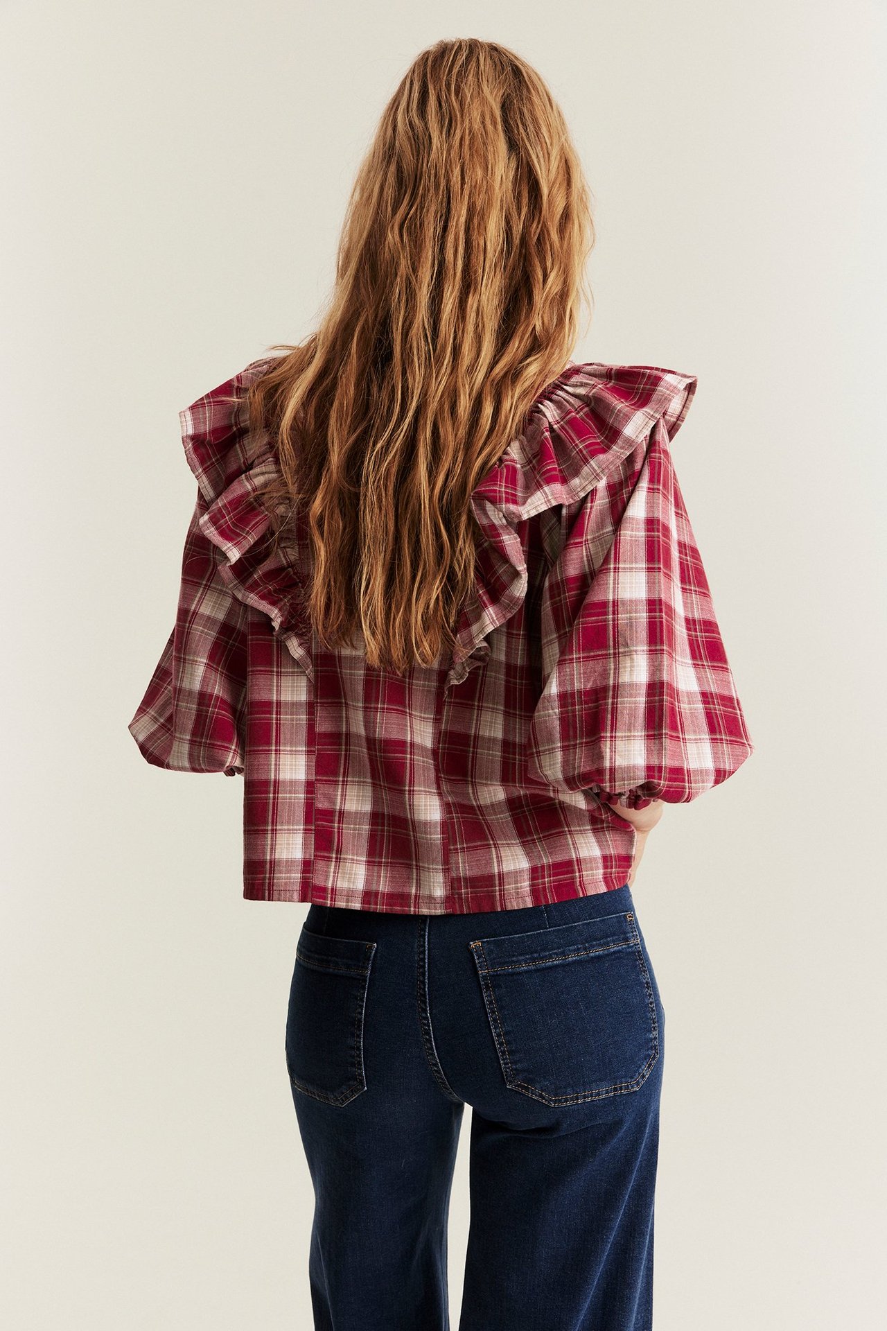 Back view of a woman wearing a red plaid cotton ruffle blouse with puff sleeves and dark blue jeans.