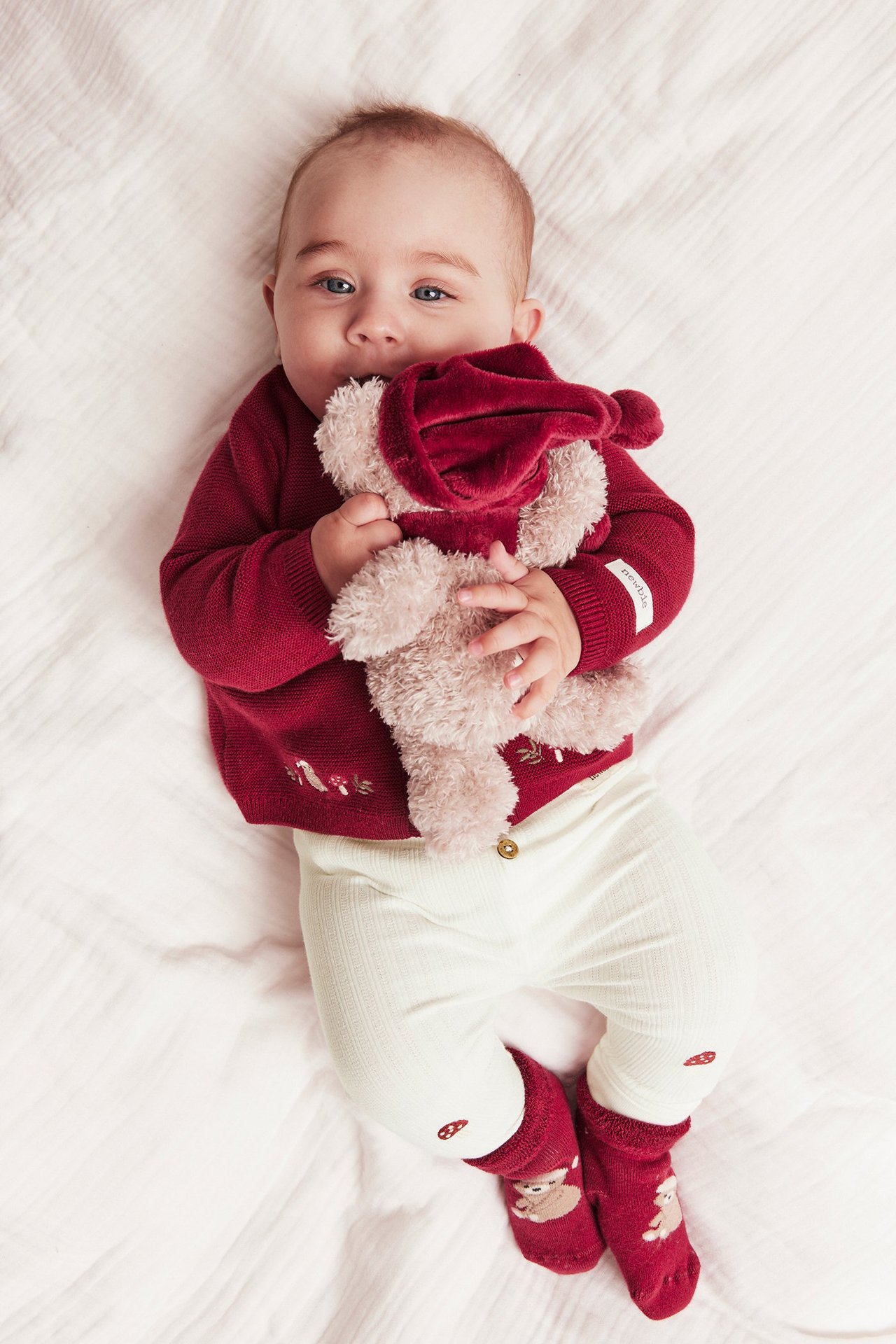 Top view: Baby wears offwhite ribbed leggings with knee embroidery, red sweater, red socks, holding plush toy.