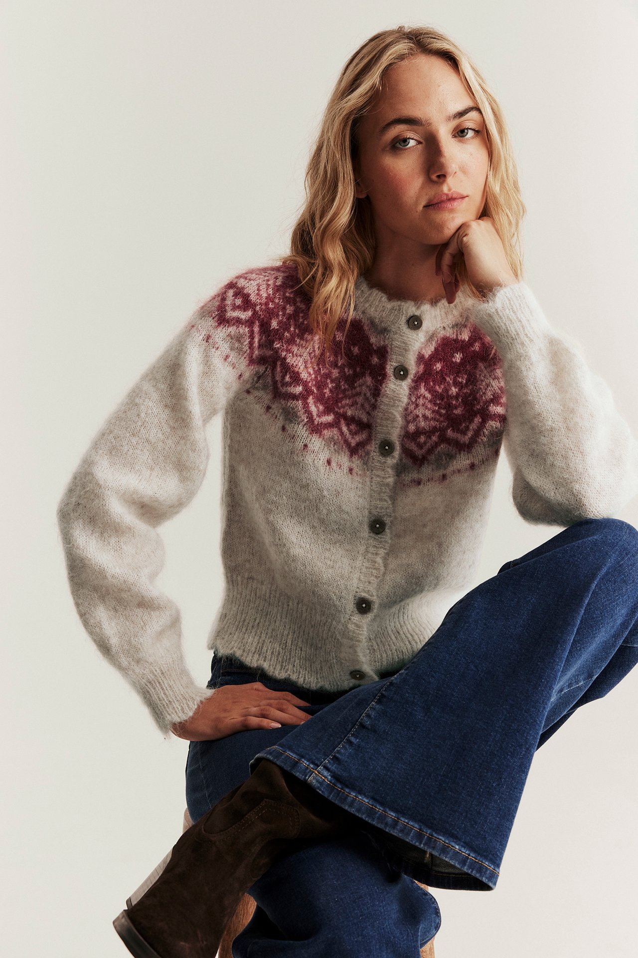 Front view of a woman wearing a beige melange fuzzy wool blend cardigan with red jacquard pattern, blue jeans, and brown boots.