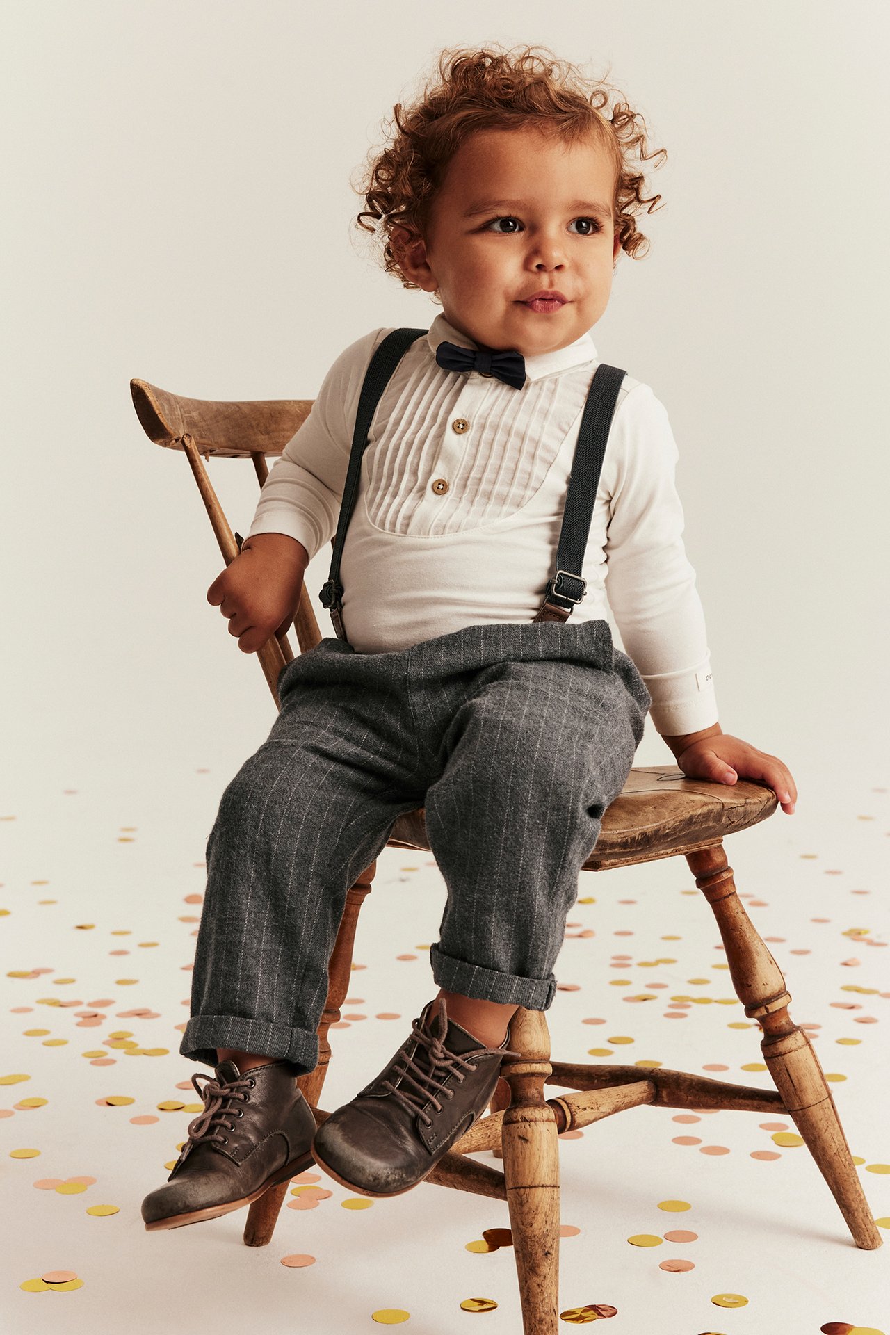 Baby Junge trägt graumelierte Nadelstreifen-Trägerhose, weißes Hemd, dunkle Fliege, braune Stiefel. Vorderansicht.