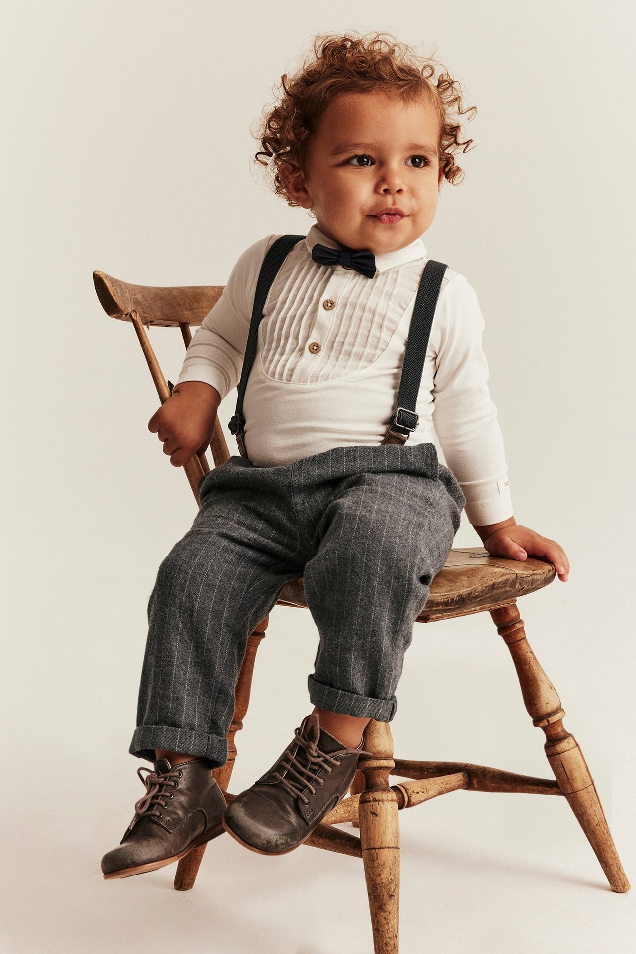 Junge in grau melierte Webhose mit Hosenträgern, weißem Hemd, schwarzem Fliege, sitzt auf Holzstuhl.